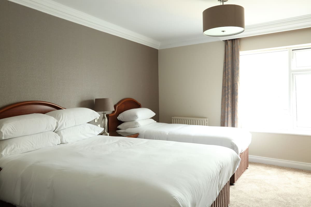 Une chambre d’hôtel avec deux lits simples, une tête de lit en bois, une table de chevet avec une lampe et une fenêtre avec des rideaux.