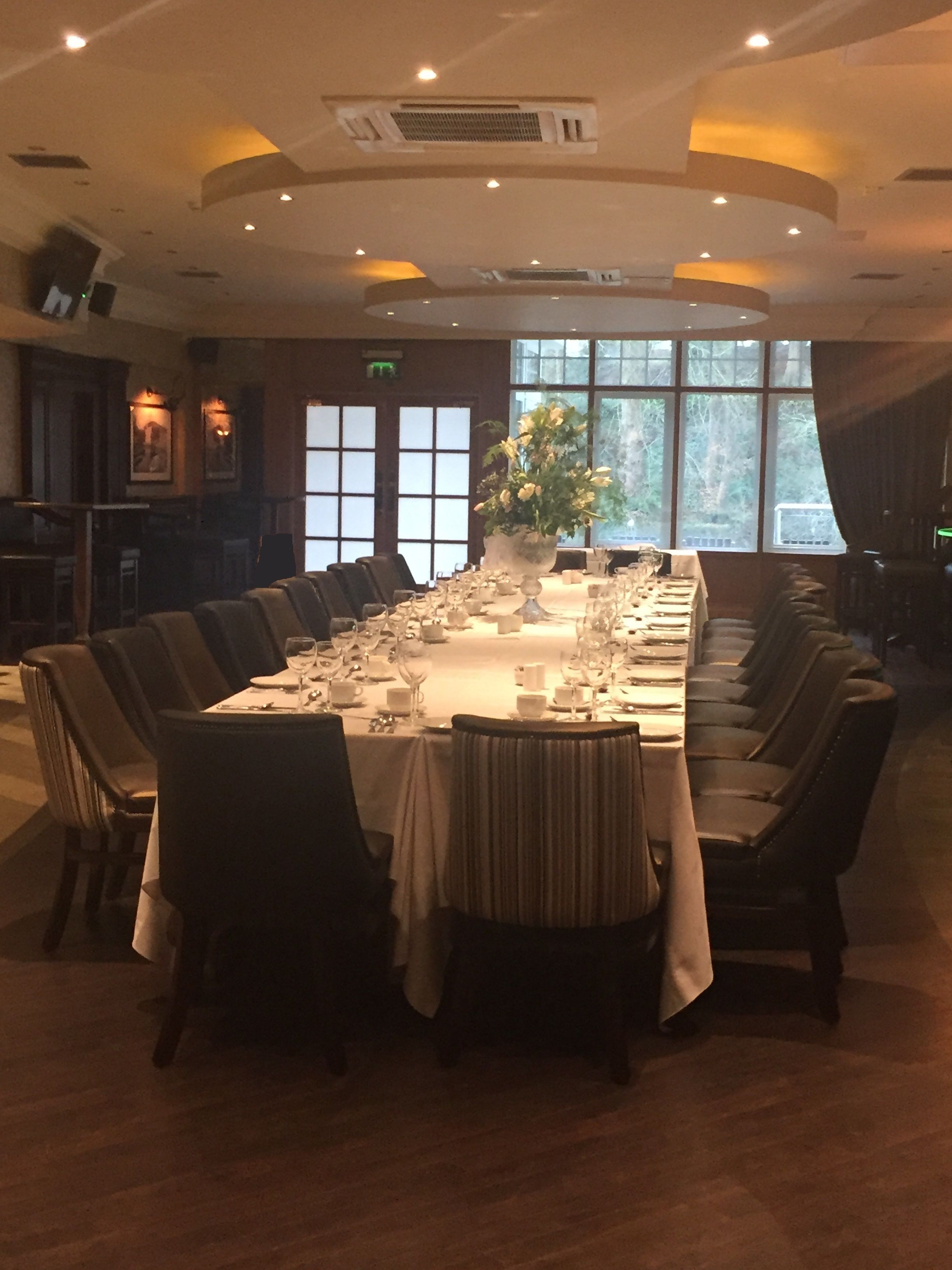 A long dining table set for a formal meal, surrounded by chairs, with a large window providing natural light and a view of trees outside.