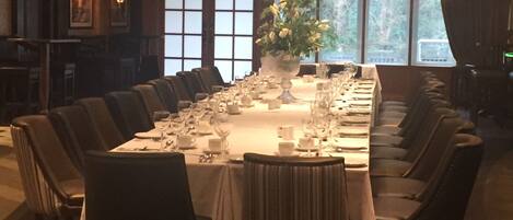 A long dining table set for a formal meal, surrounded by chairs, with a large window providing natural light and a view of trees outside.