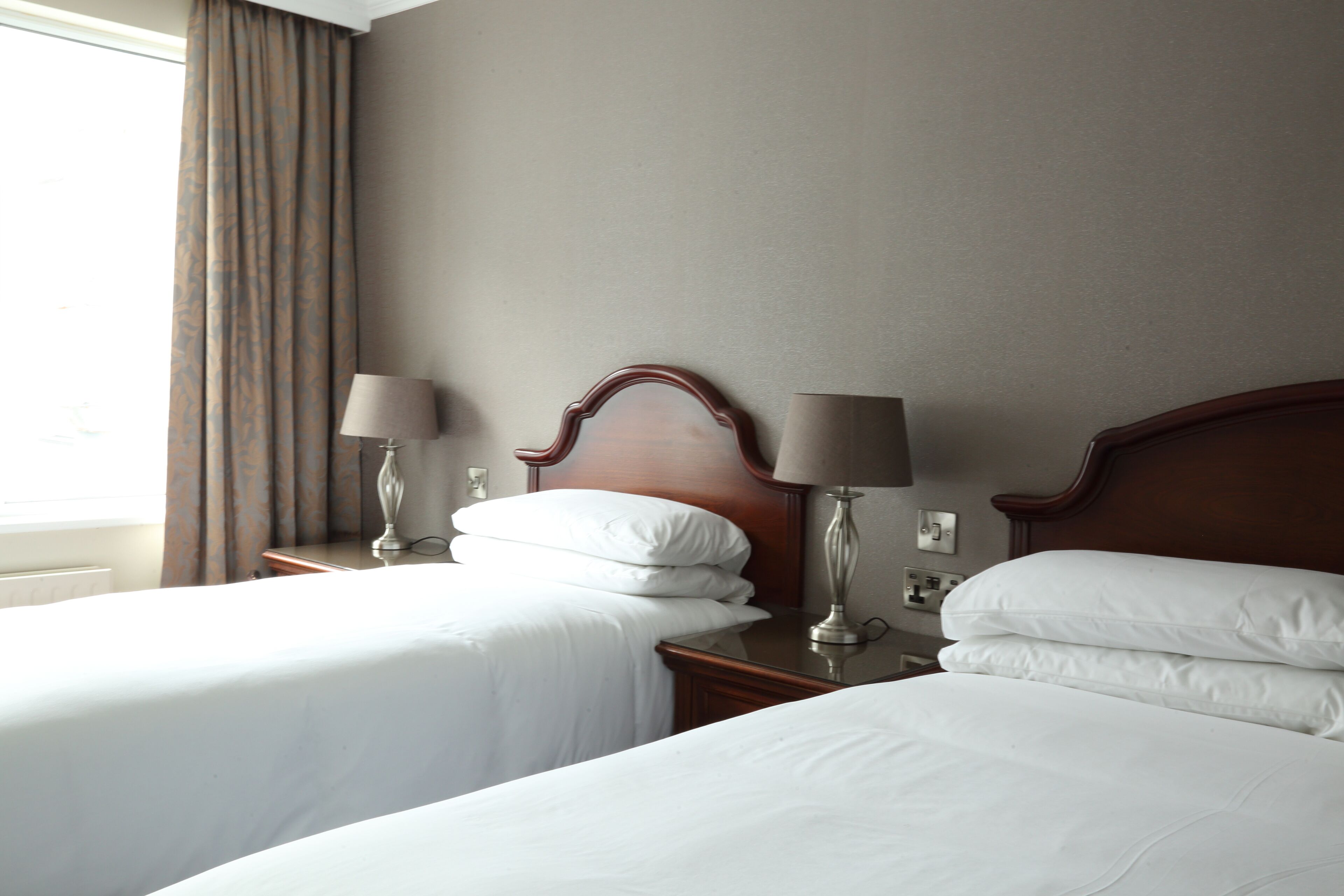 Une chambre d’hôtel avec deux lits simples, chacun doté d’une tête de lit en bois, d’une table de chevet avec une lampe et d’une fenêtre avec des rideaux.