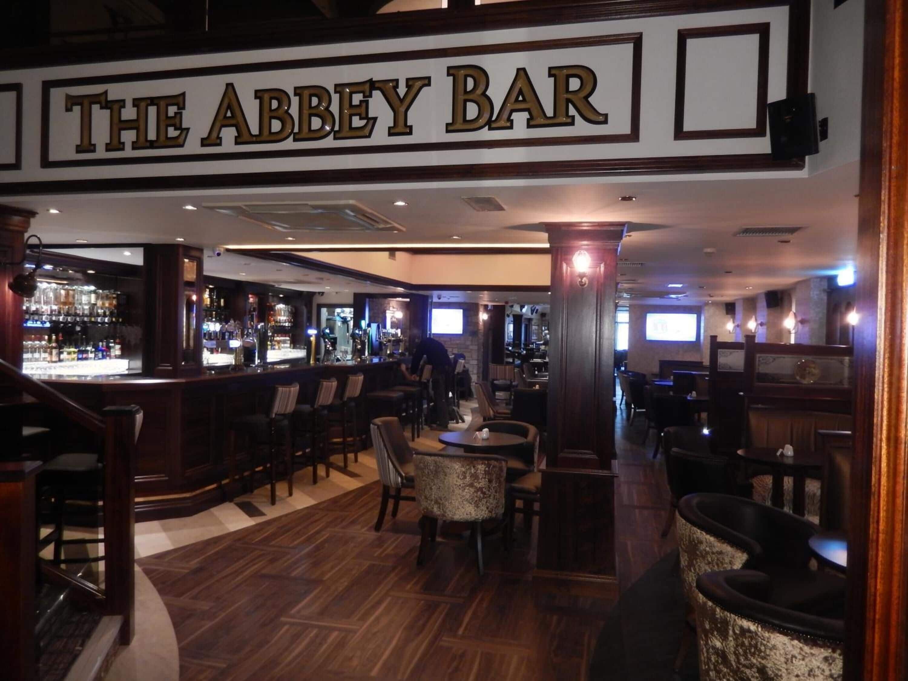Un bar con un letrero que dice "THE ABBEY BAR". Tiene suelo de madera, taburetes de bar y mesas.
