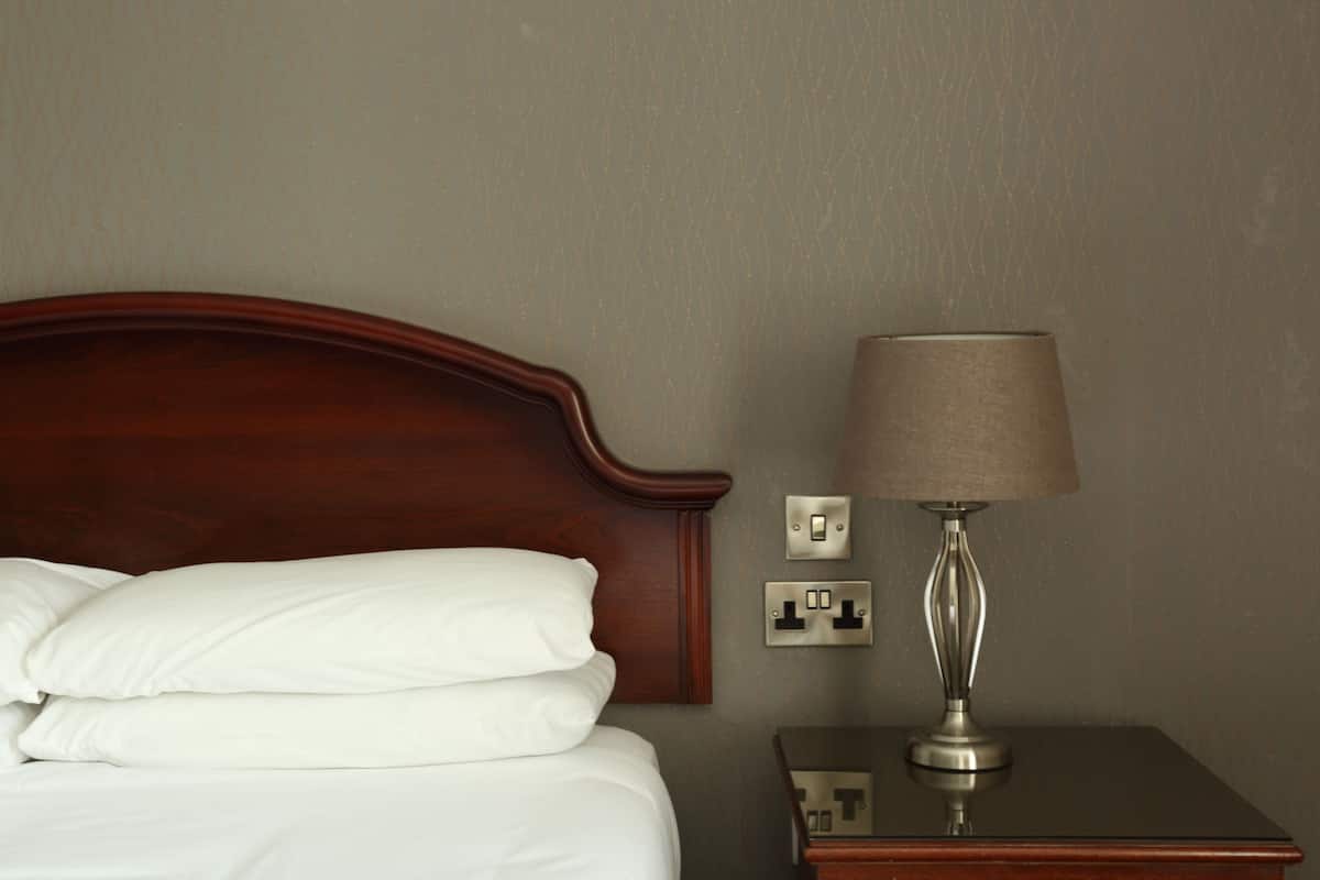 Un lit bien fait, avec du linge de lit blanc et une tête de lit en bois, une table de chevet avec une lampe et une prise de courant sur le mur.