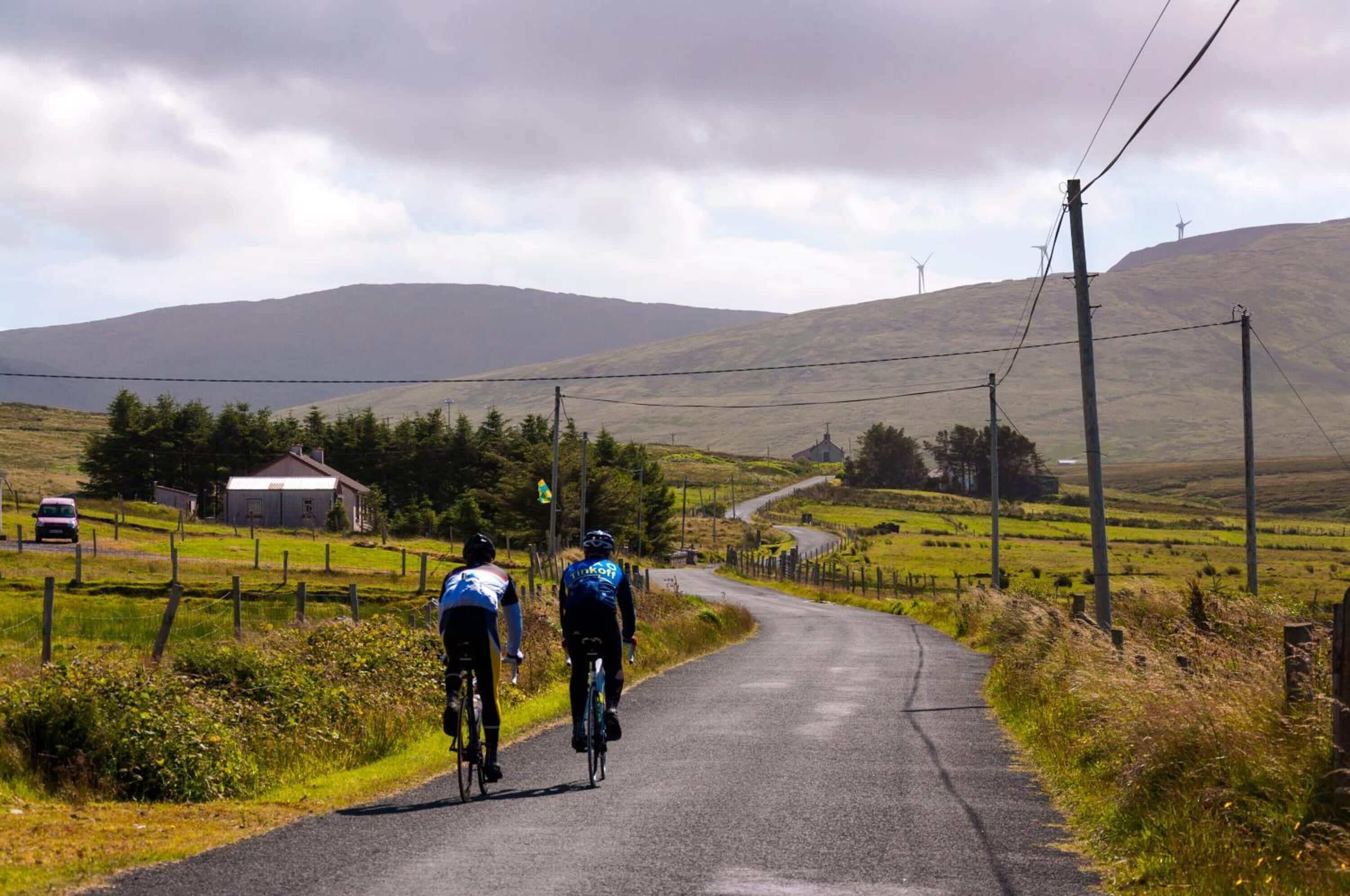 A rural road with cyclists, a farmhouse, and wind turbines in the background.