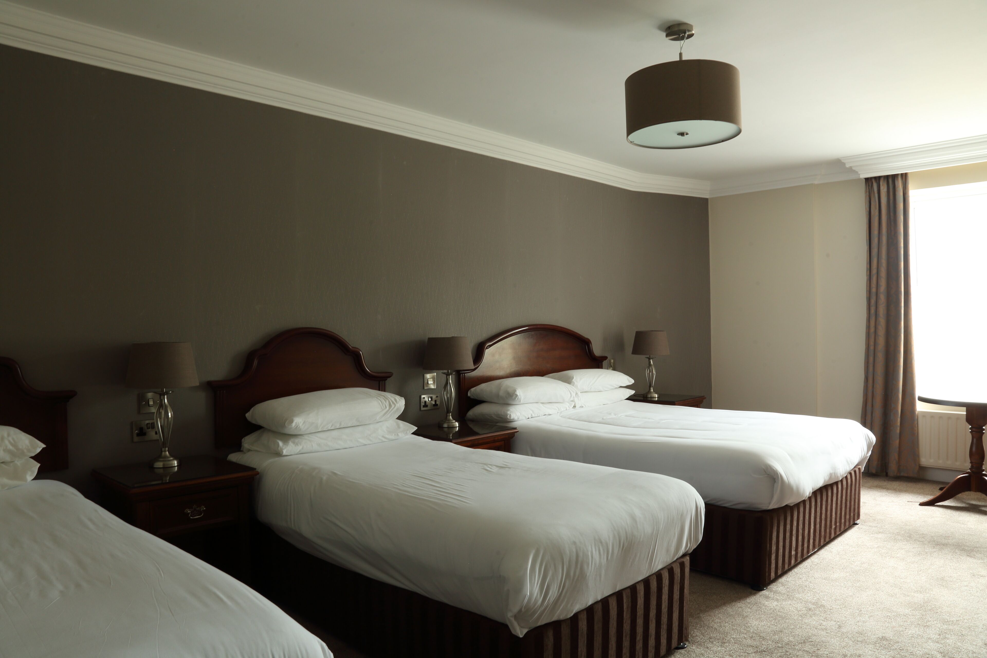 A hotel room with two single beds, wooden headboards, bedside tables with lamps, and a large window with curtains.