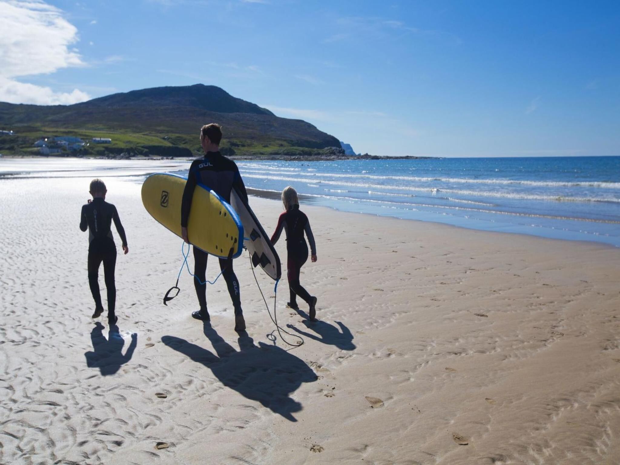 Escena de playa con una persona llevando una tabla de surf y dos niños caminando sobre la arena.