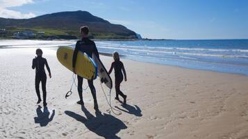 Escena de playa con una persona llevando una tabla de surf y dos niños caminando sobre la arena.