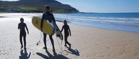 Escena de playa con una persona llevando una tabla de surf y dos niños caminando sobre la arena.