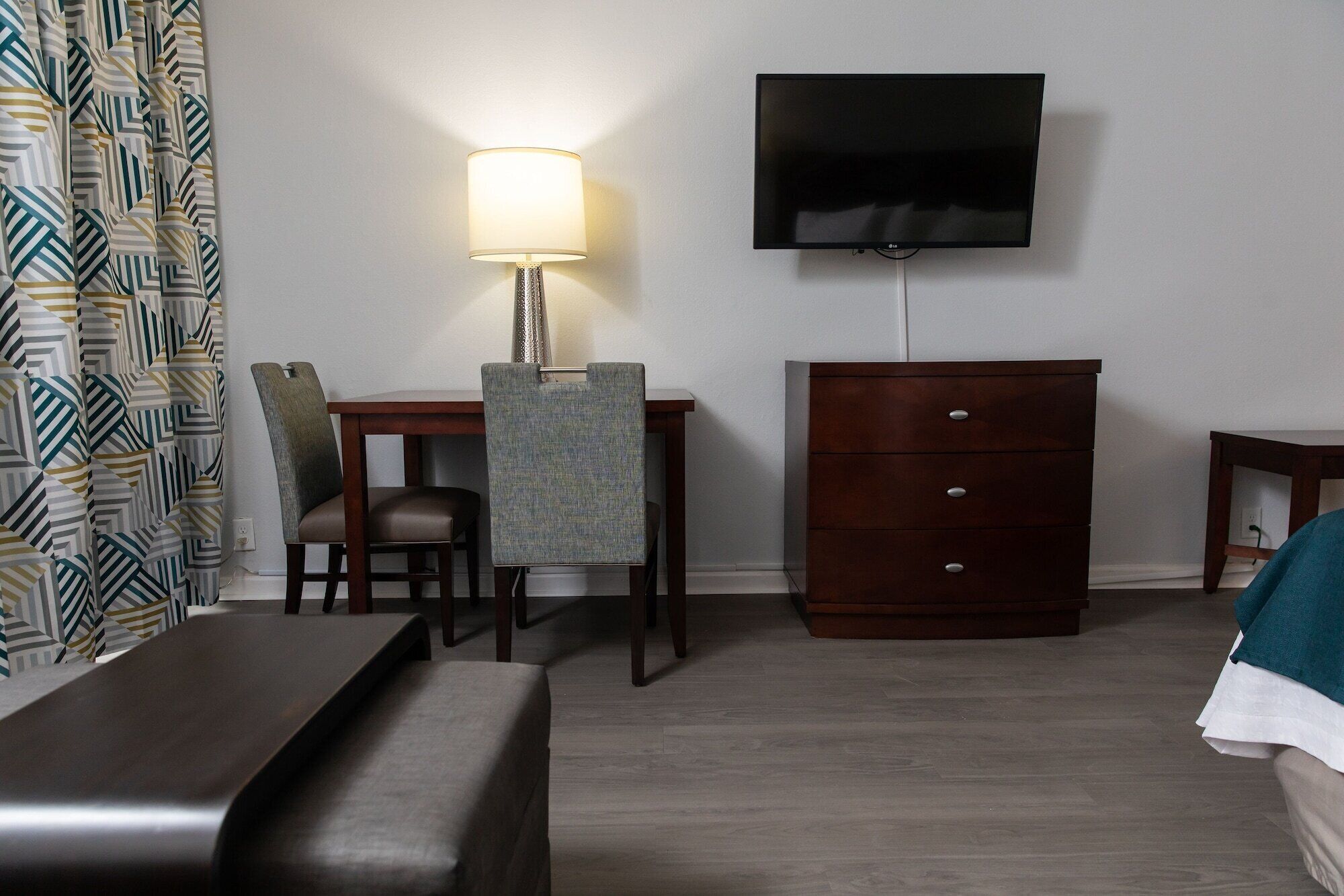 A hotel room with a bed, a desk with a lamp, a TV, a dresser, and a patterned curtain.