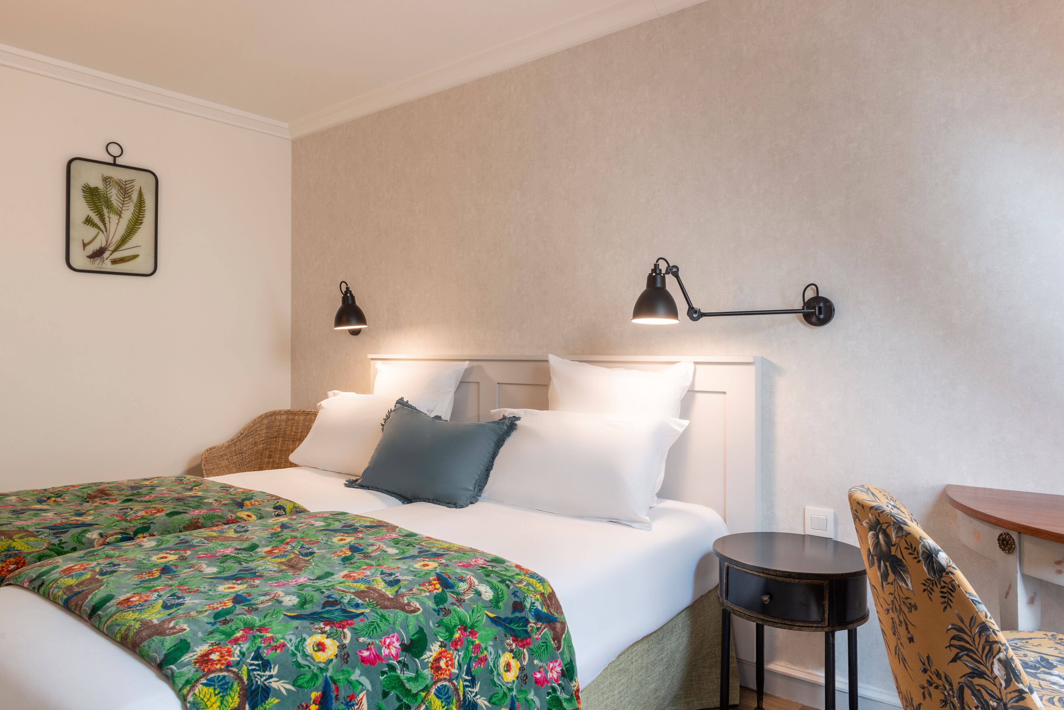 A neatly made bed with a floral bedspread, two bedside lamps, a small round table, and a chair with a floral pattern.