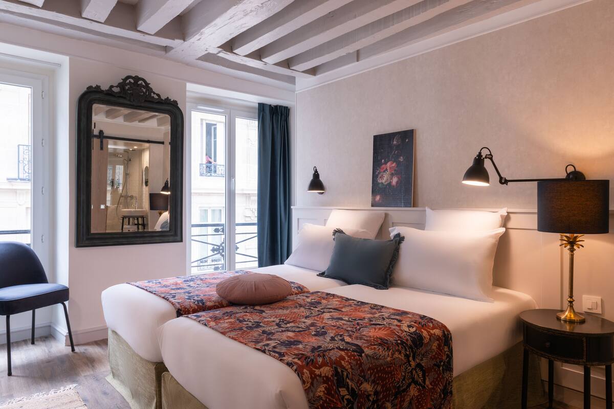 A hotel room with a bed, a chair, a desk, a mirror, and a window with curtains.