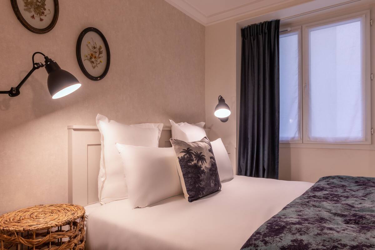 A neatly made bed with white and patterned pillows, a wicker side table, and two wall-mounted lamps.