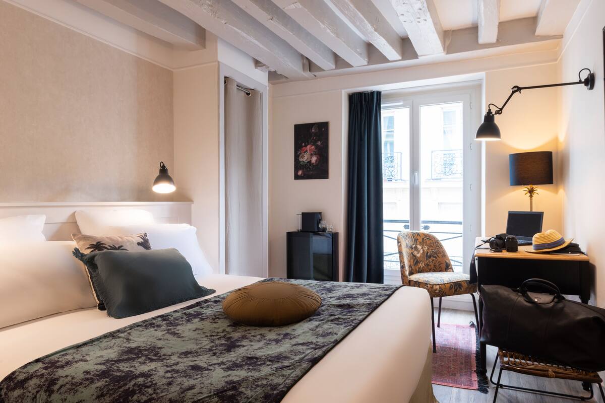 A hotel room with a bed, a desk with a laptop, a chair, and a view of the outside.