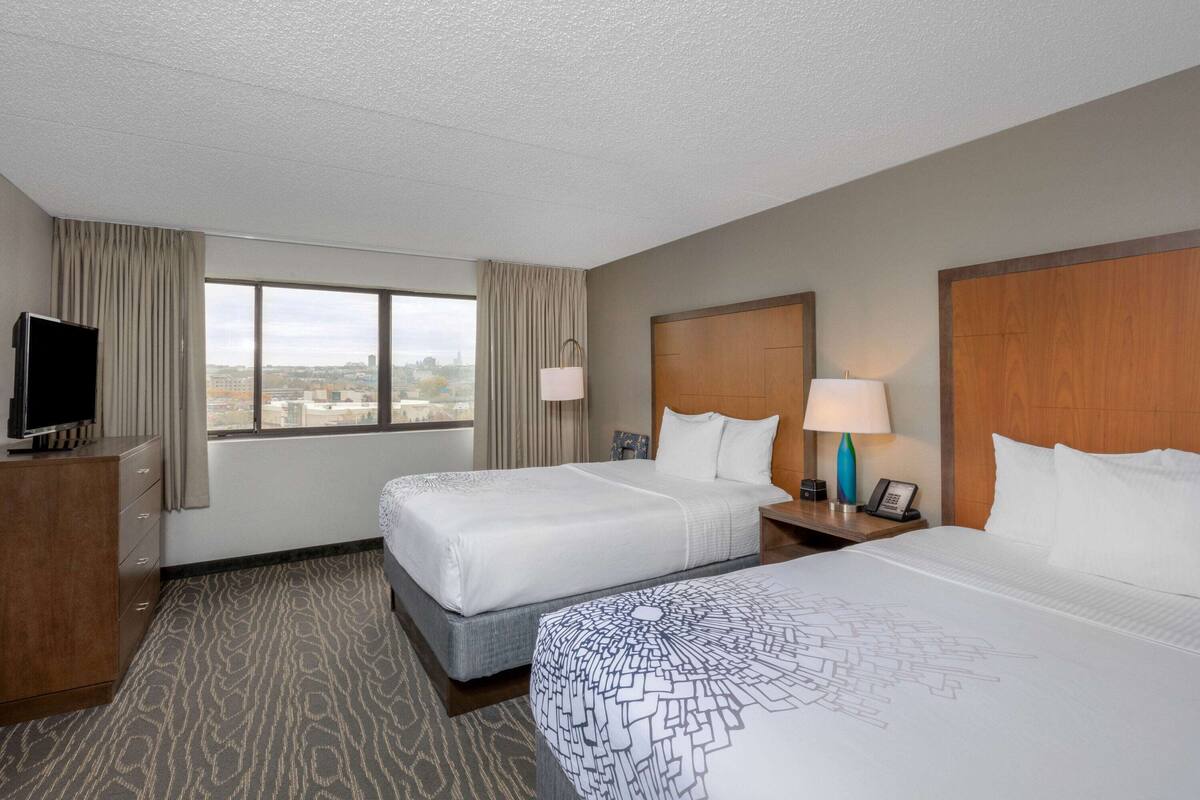 Ein Hotelzimmer mit zwei Betten, einem Fernseher, einem Schreibtisch und einem Fenster mit Blick auf die Stadt.