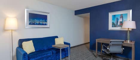 A hotel room with a blue velvet sofa, a small side table, a desk with a chair, and two framed pictures on the walls.