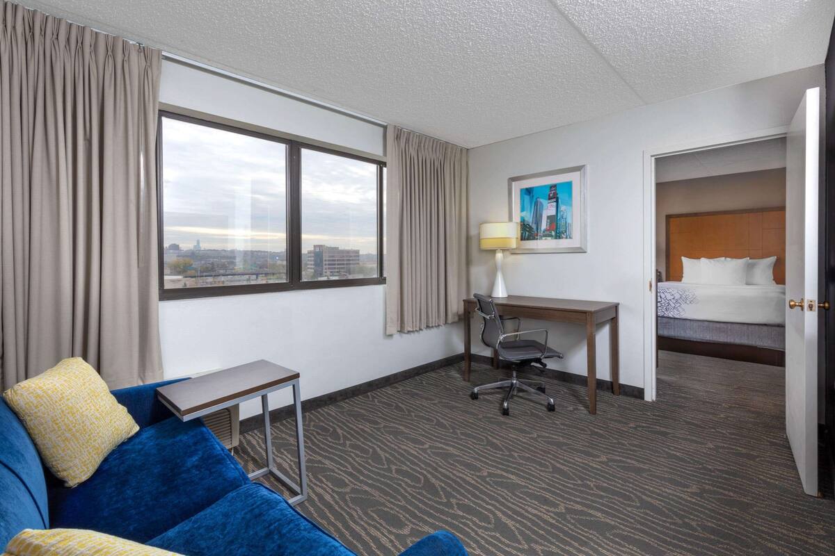 Ein Hotelzimmer mit großem Fenster, Schreibtisch mit Lampe, blauem Sofa und einem Bett.