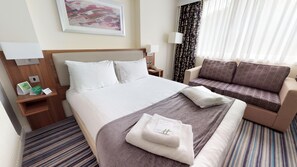 A hotel room with a bed, a sofa, a bedside table with a lamp, a telephone, and a framed picture on the wall.