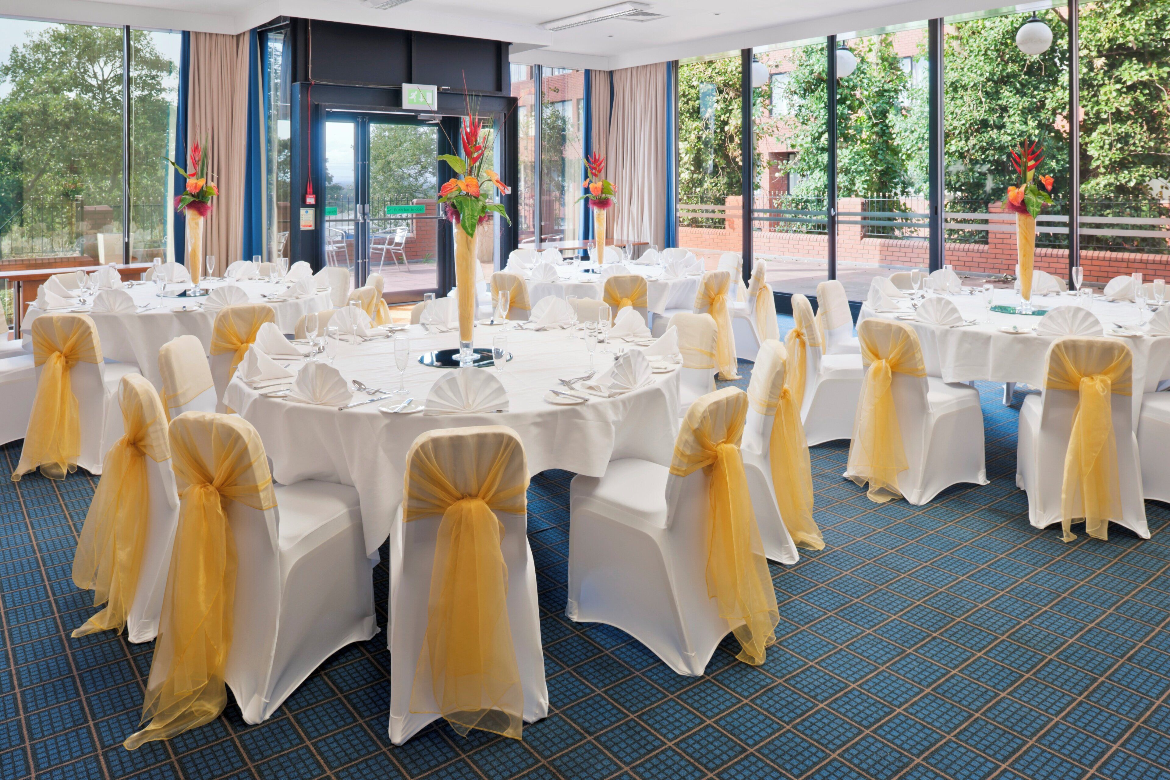 A banquet hall with round tables, white chairs with yellow sashes, and large windows with a view of greenery.