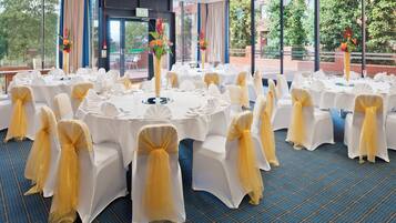 A banquet hall with round tables, white chairs with yellow sashes, and large windows with a view of greenery.