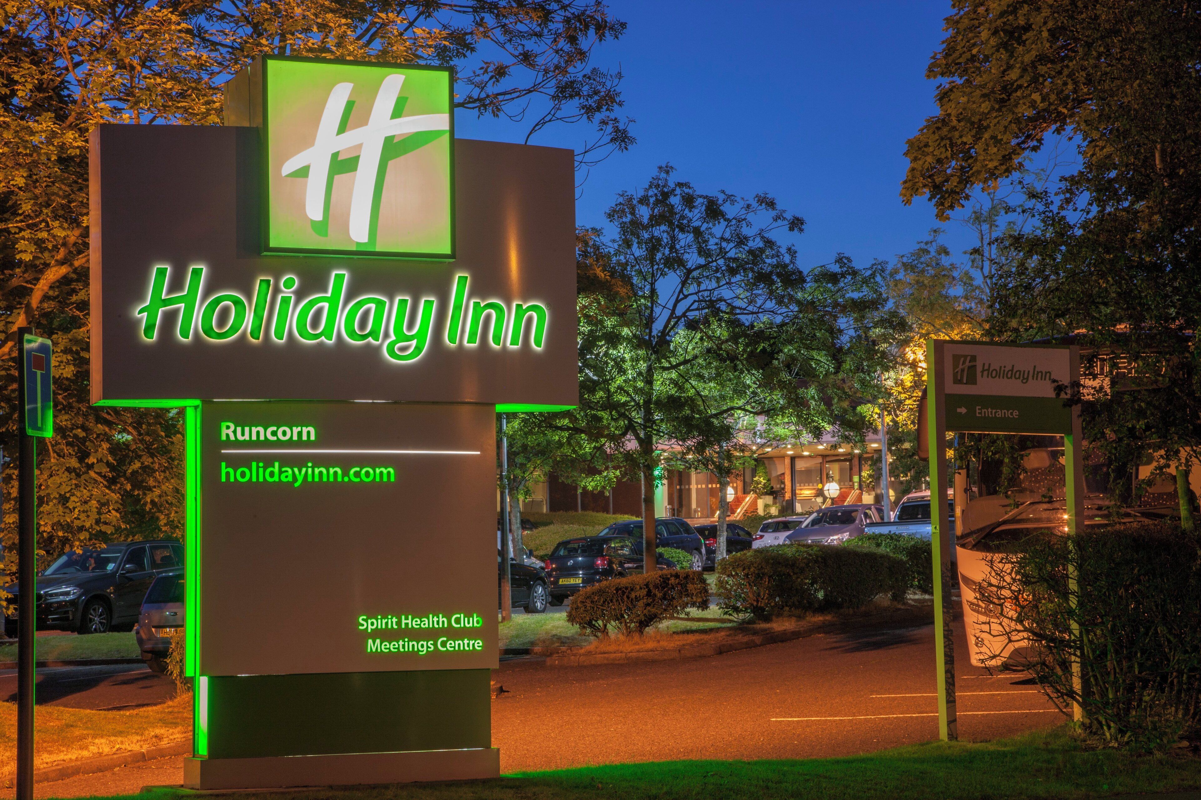 A Holiday Inn sign with a green neon light, a parking lot with cars, and a building in the background.