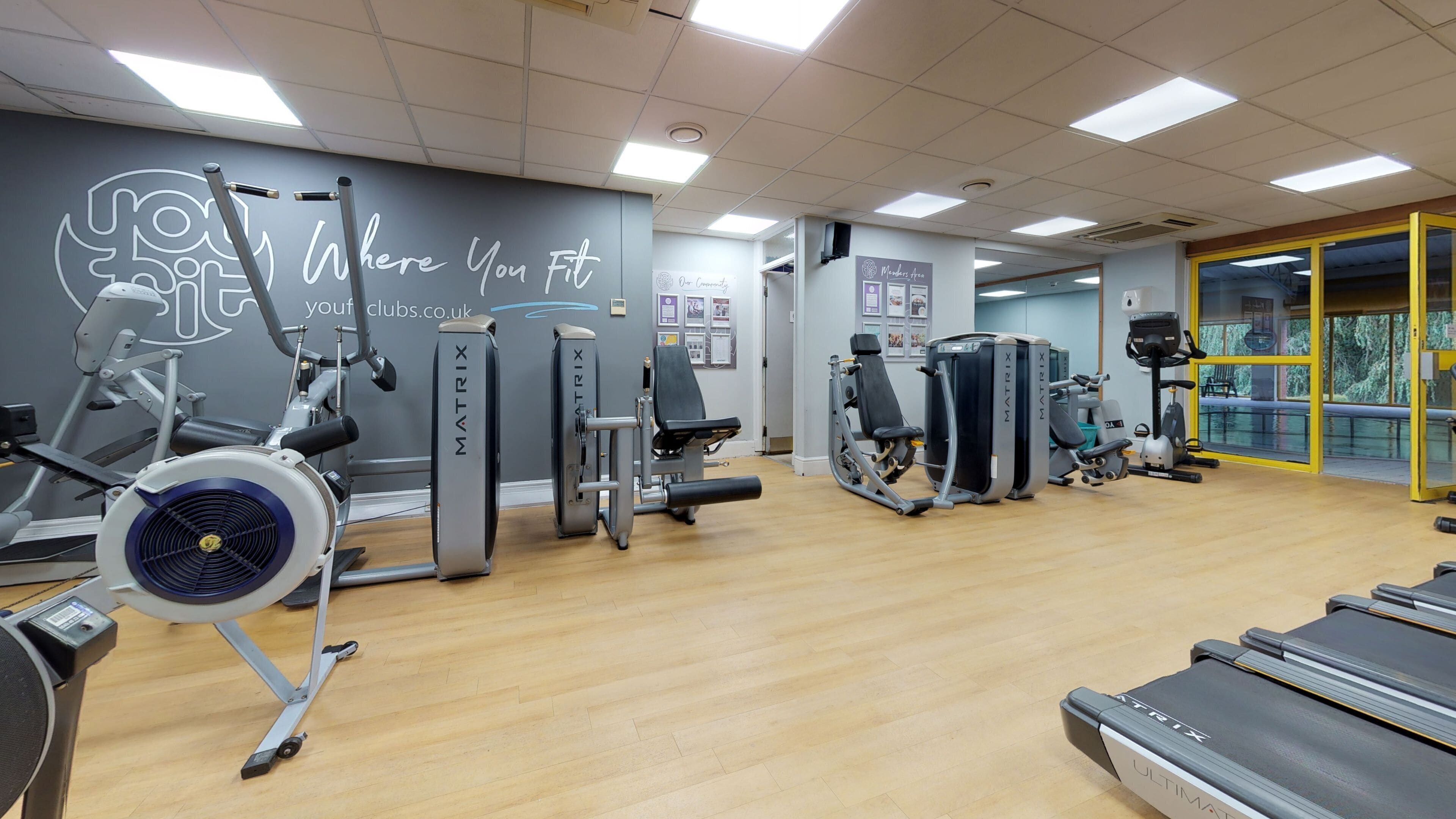 Ein Fitnessraum mit unterschiedlichen Trainingsgeräten, einer Wand mit motivierenden Texten und Blick auf Bäume durch eine Glastür.
