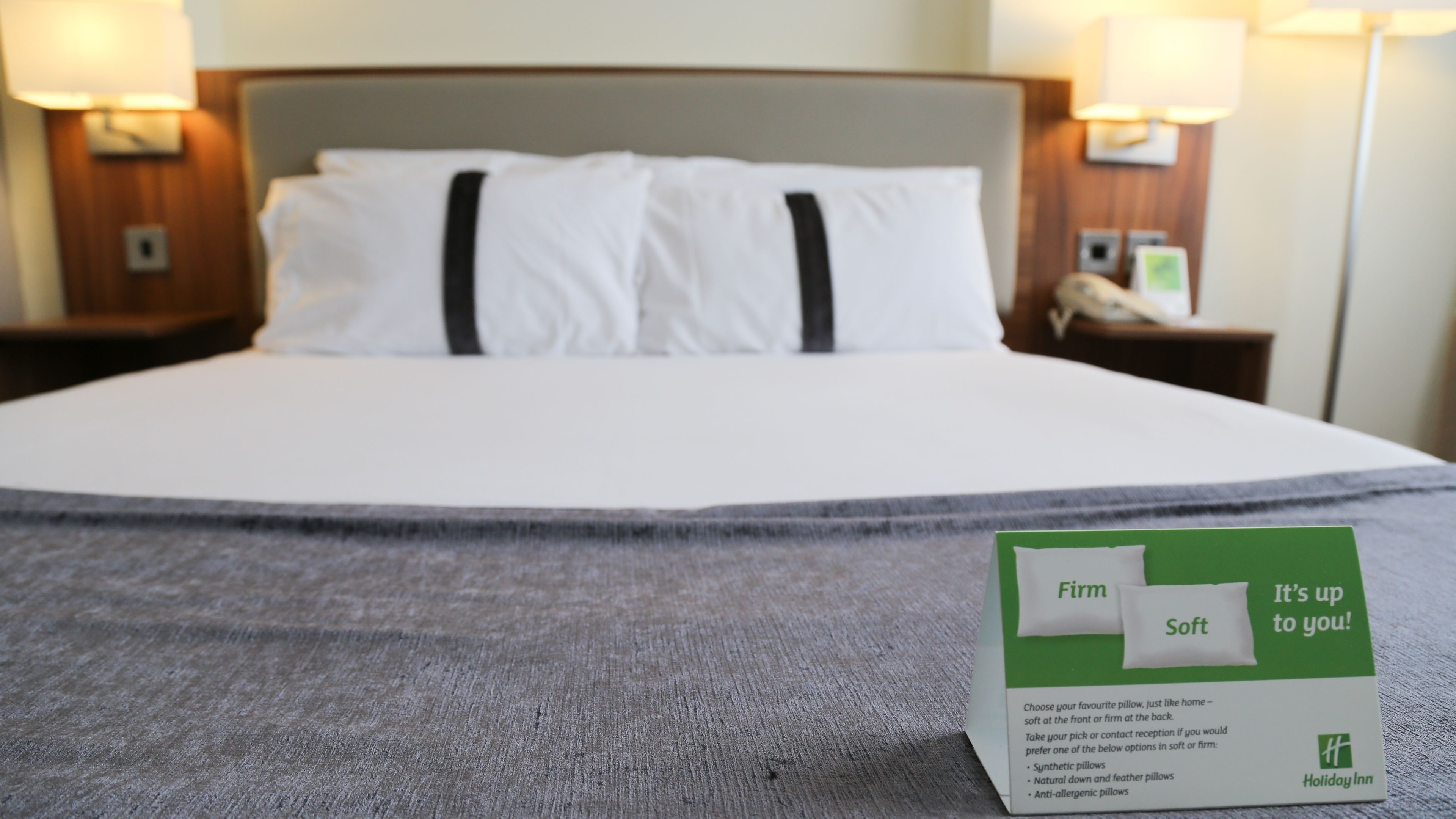 Cama bem arrumada com lençóis brancos e duas almofadas pretas, um cartão de escolha de almofadas verde e branco na cama, e uma mesa de cabeceira com abajur e telefone.