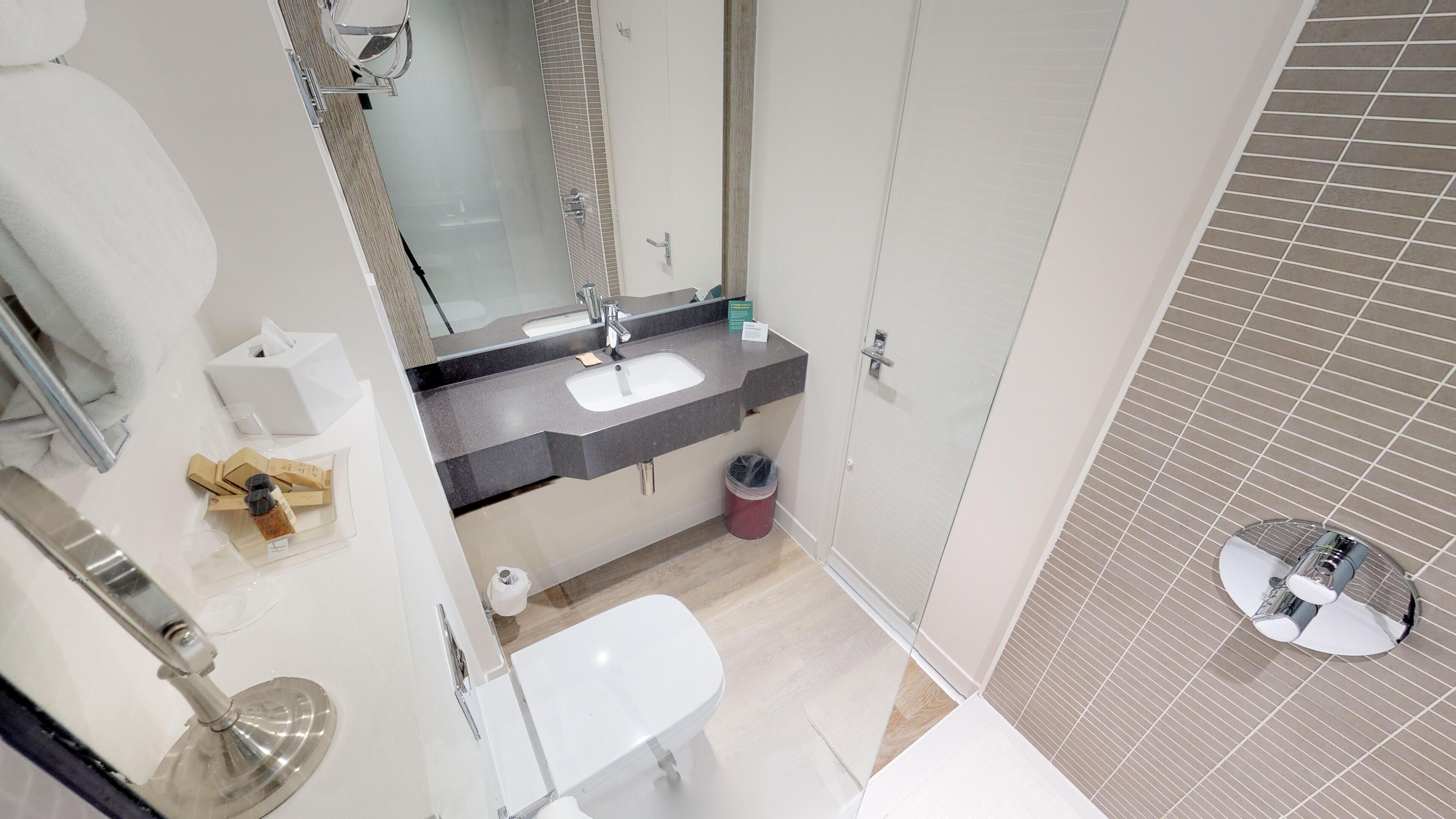 A modern bathroom with a white sink, mirror, and toilet. There is a trash can and a small window with blinds.
