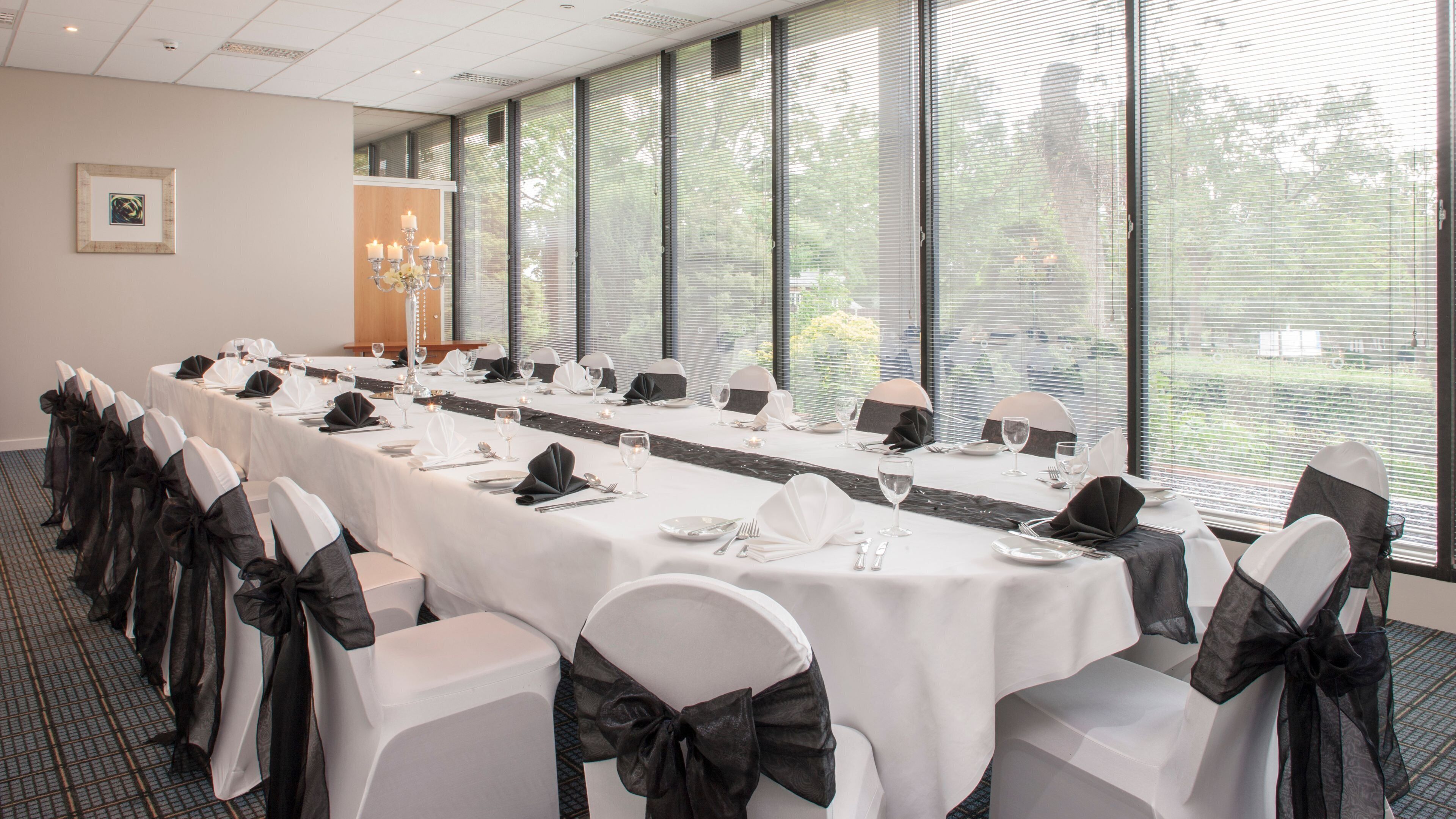 Un salón de conferencias con una larga mesa dispuesta para una reunión, ventanas amplias y un candelabro.
