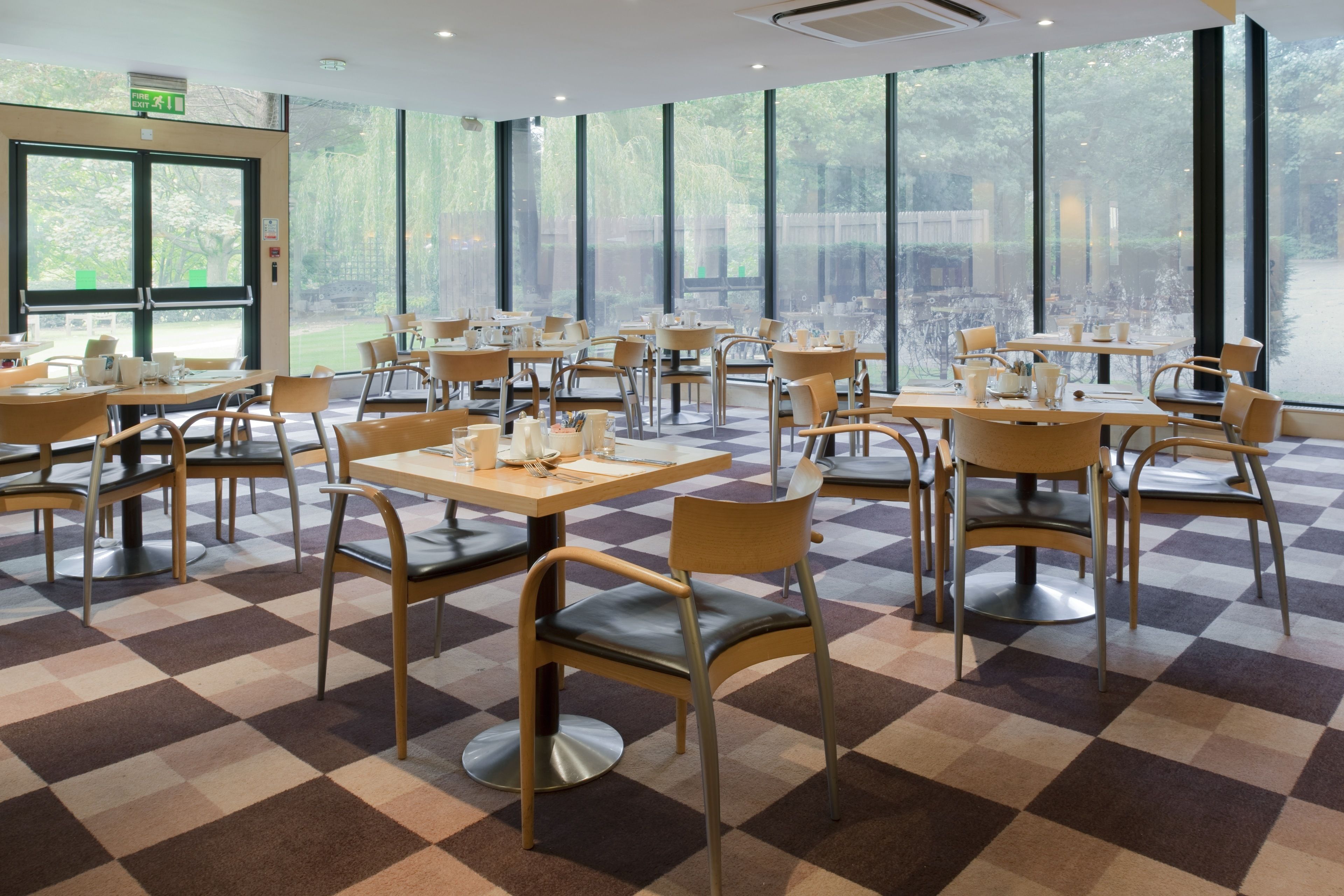 A spacious cafe with checkered flooring, wooden tables, and chairs. Large windows offer a view of the outdoors.
