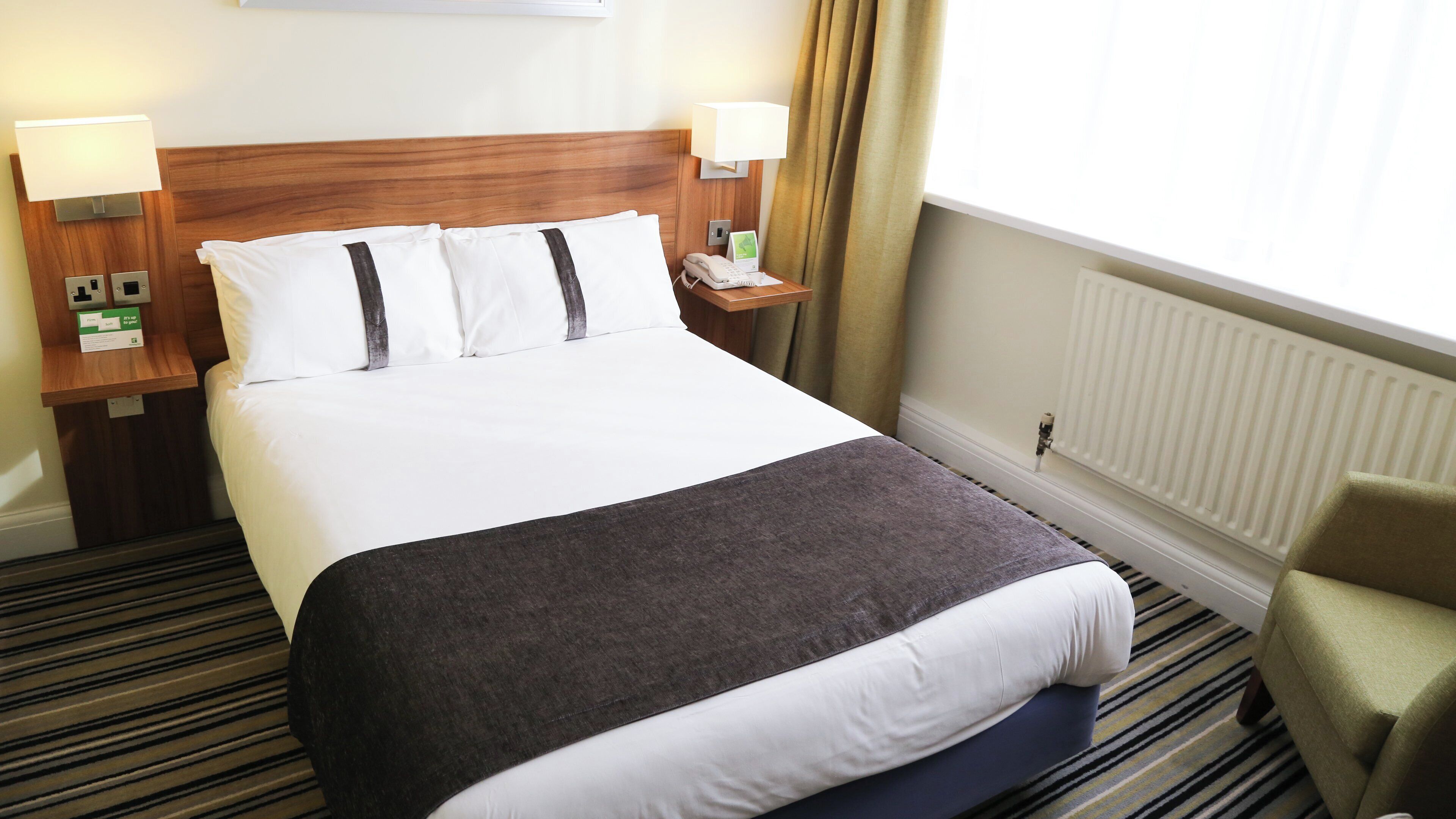 A hotel room with a large bed, two bedside tables with lamps, a telephone, a thermostat, and a window with curtains.