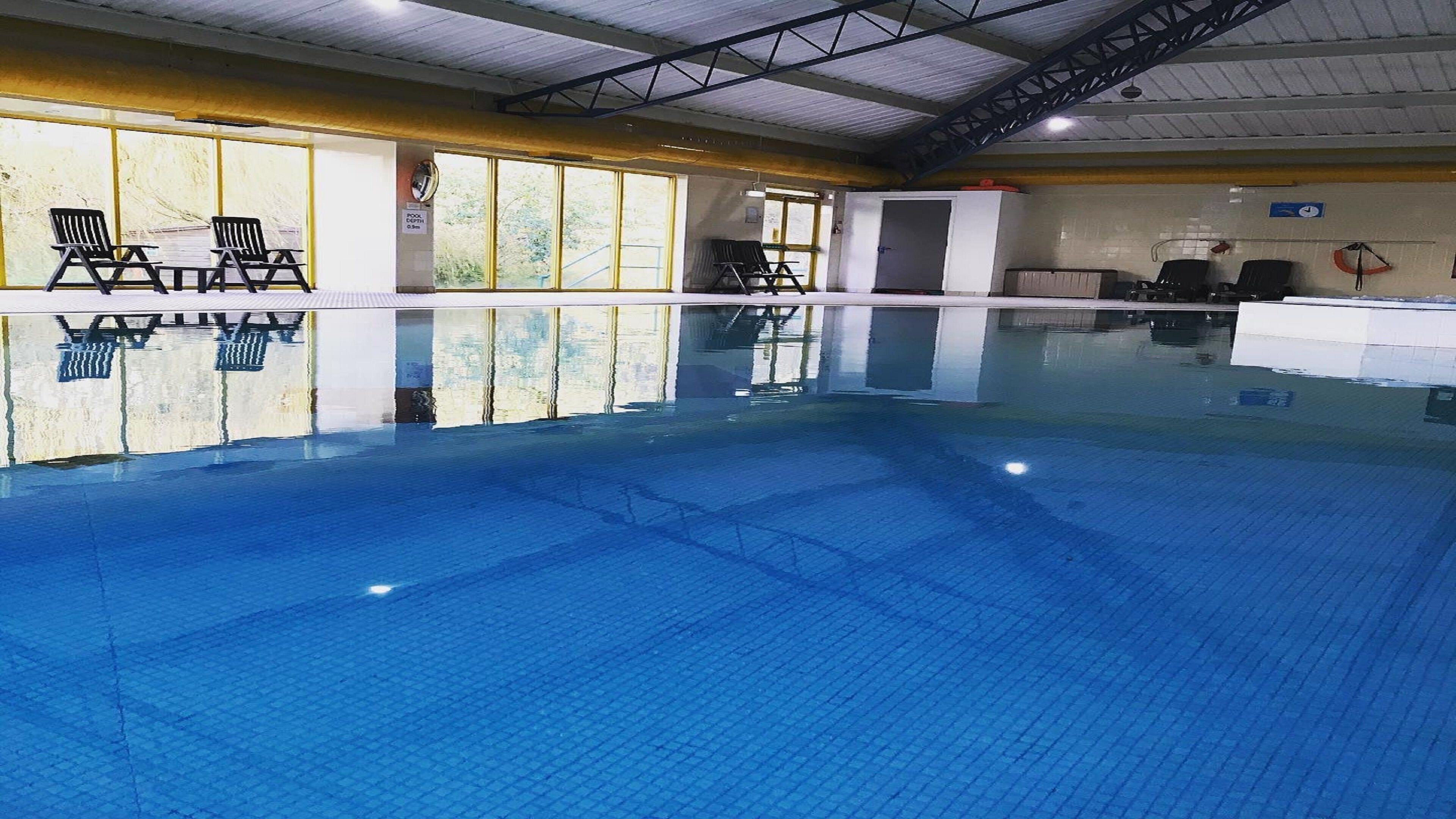 An indoor swimming pool with clear water, a tiled floor, and a pool depth sign. There are lounge chairs along the poolside and a lifeguard chair.