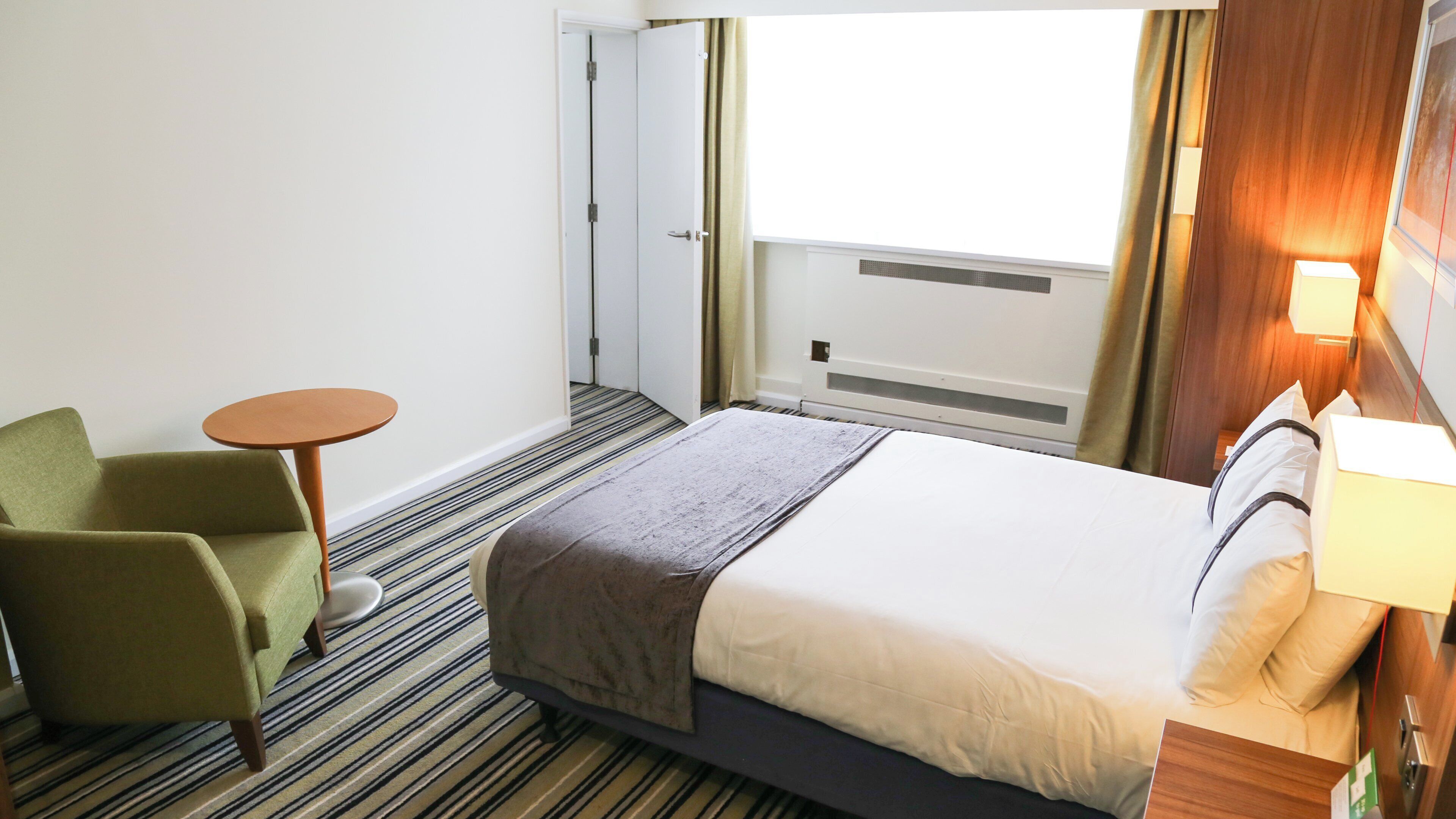 A hotel room with a bed, a chair, a small table, a window with curtains, and a bedside lamp.