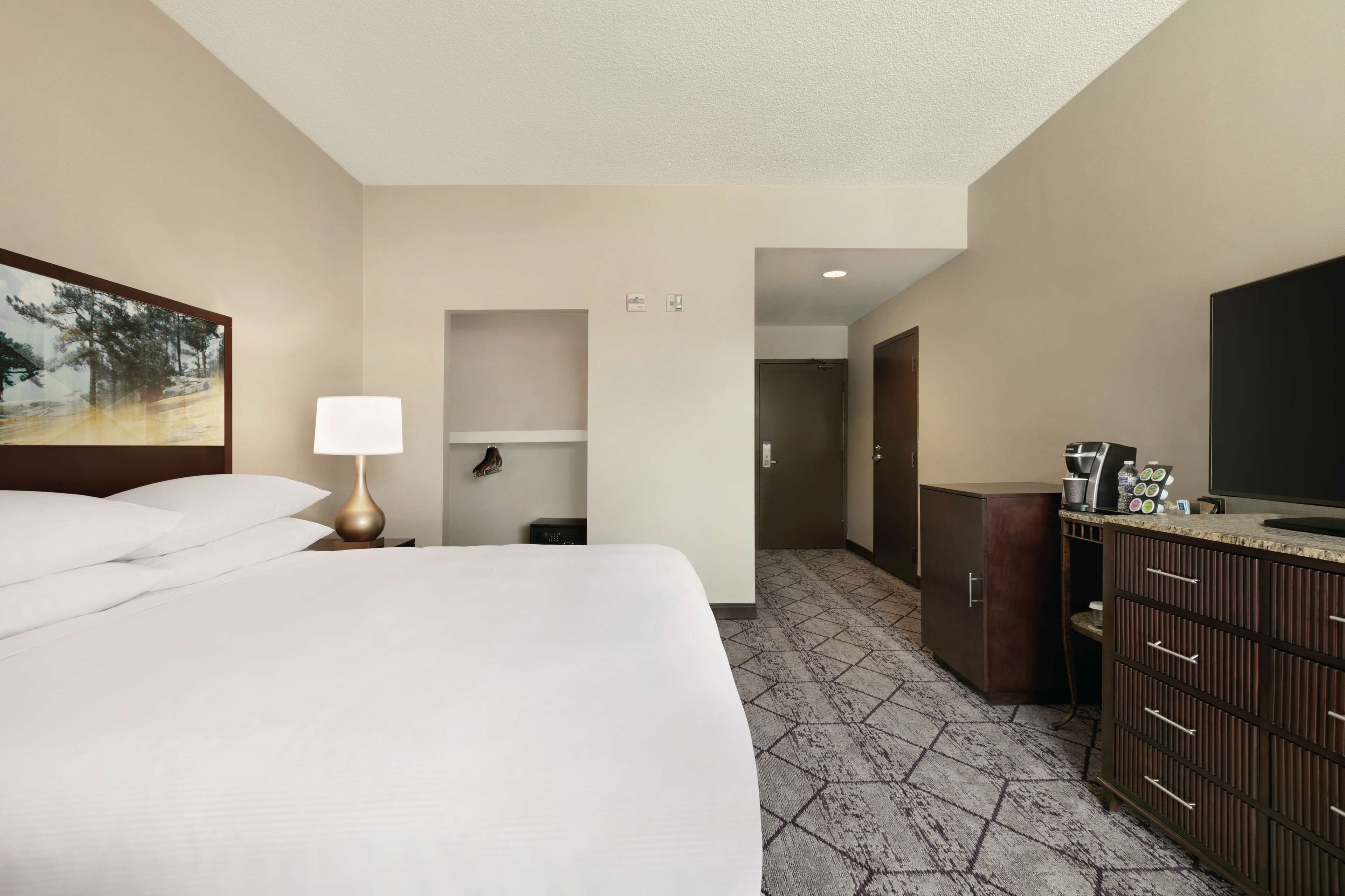 A hotel room with a large bed, a bedside lamp, a painting on the wall, a small shelf, a wardrobe, and a television.