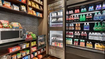 Zona de comida y bebidas de servicio propio, con microondas, refrigeradores y estantes llenos de aperitivos, bebidas y salsas.