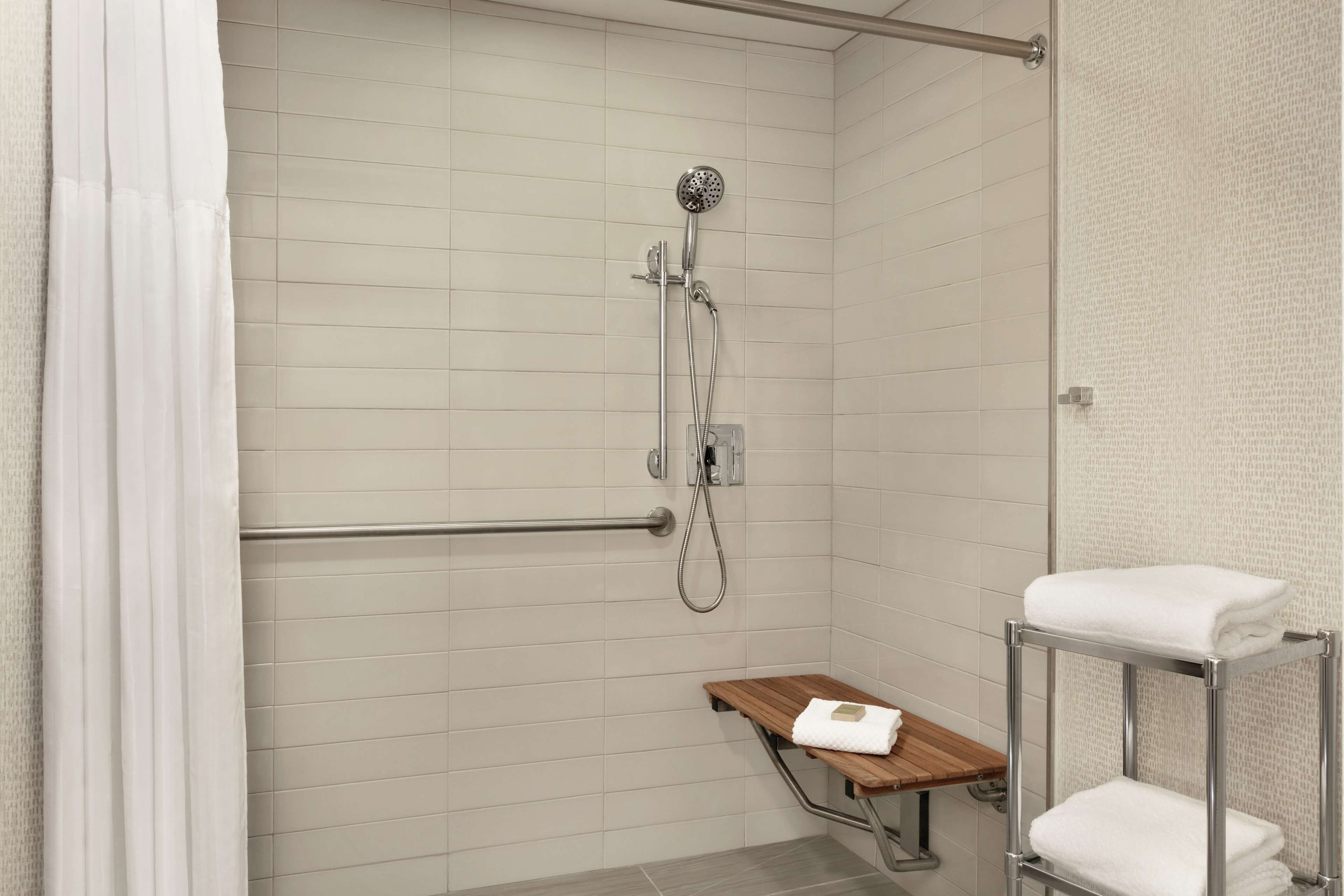 Zona de ducha con cabezal de ducha de mano, barra de apoyo y repisa de madera con una toalla.