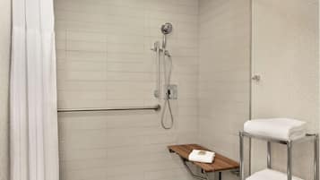 Zona de ducha con cabezal de ducha de mano, barra de apoyo y repisa de madera con una toalla.