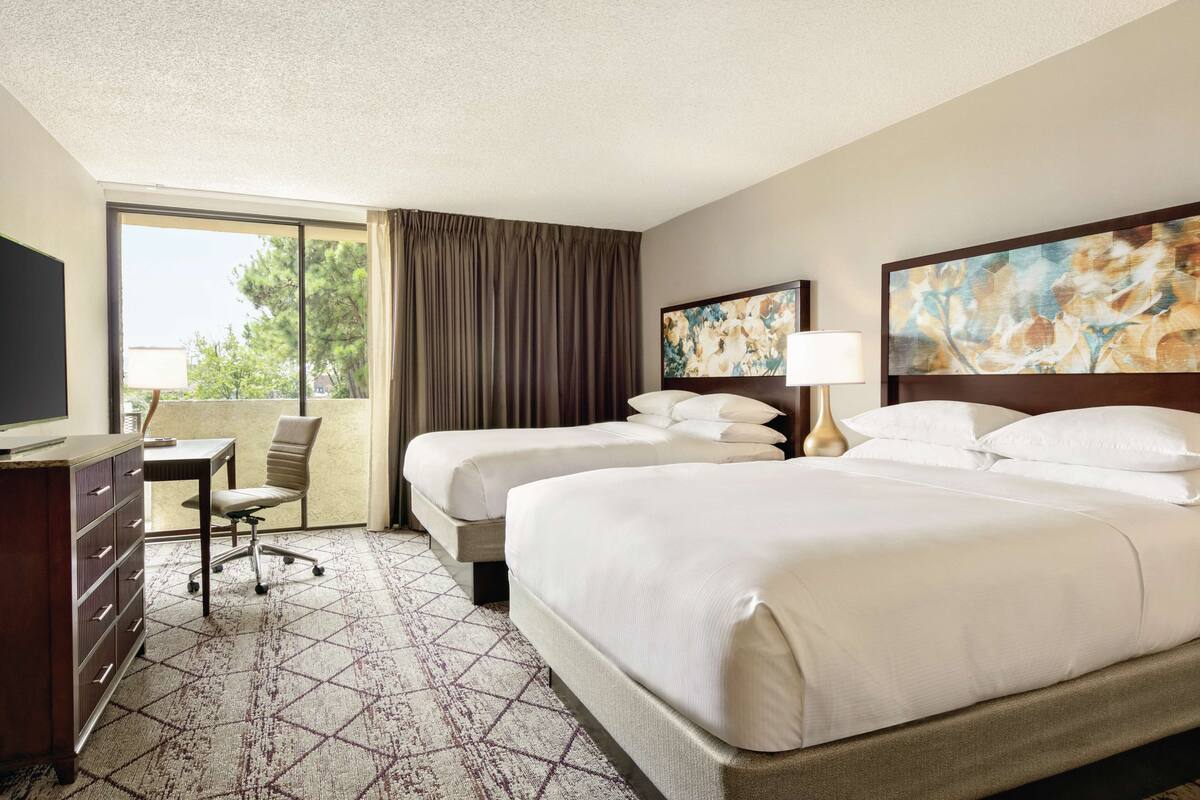 A hotel room with two beds, a desk, a chair, and a large window with curtains.