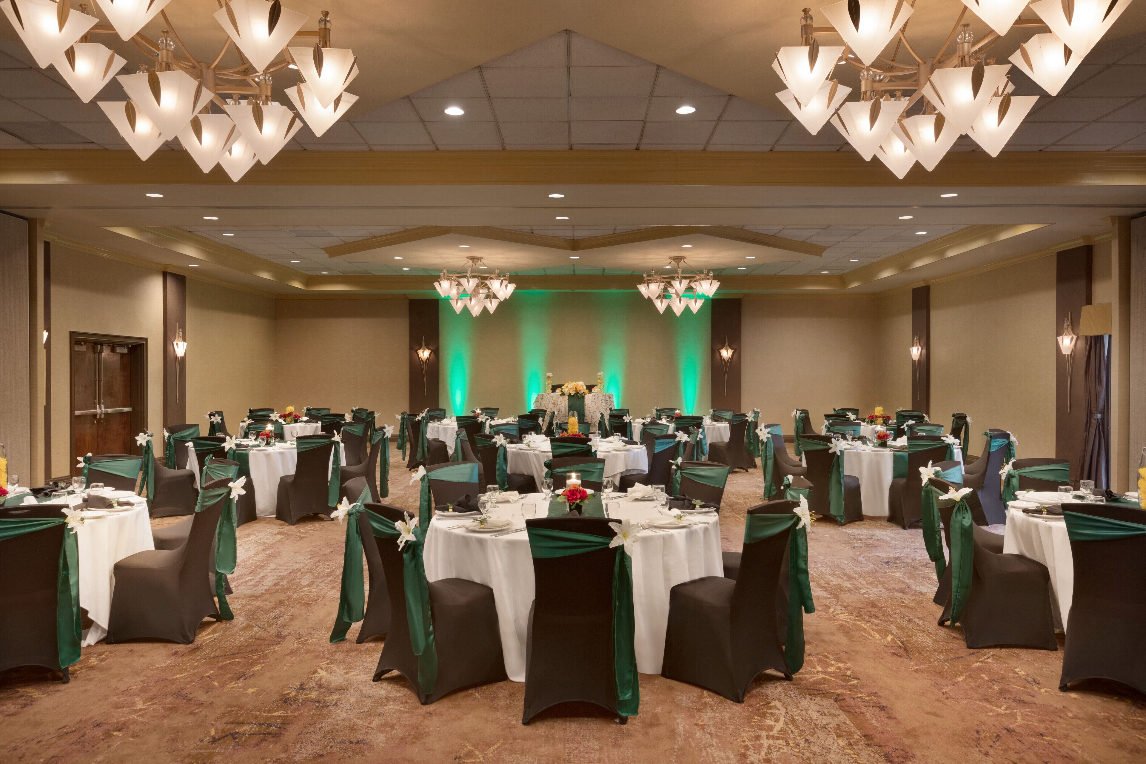 A banquet hall with round tables, green chair covers, and chandeliers.