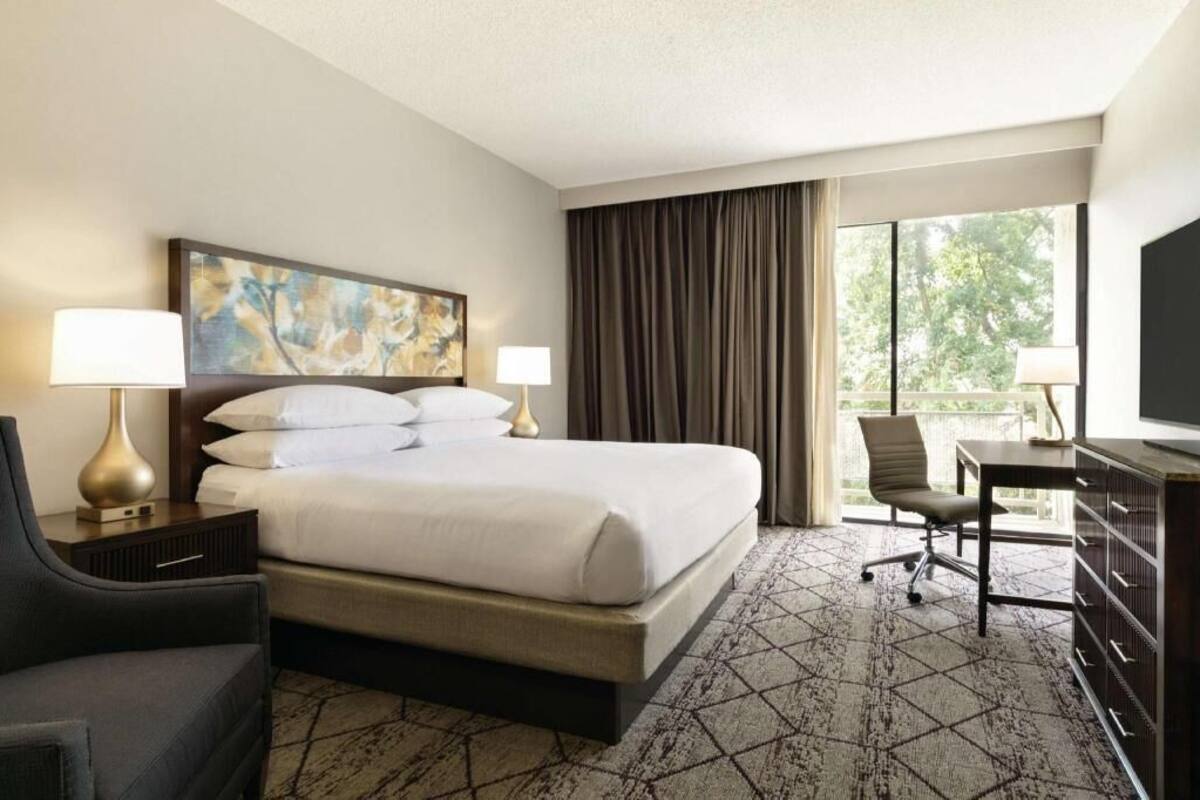 A hotel room with a bed, two bedside lamps, a desk with a chair, a television, and a large window with curtains.