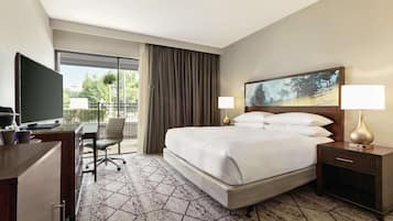 A hotel room with a large bed, a desk with a TV, a chair, a nightstand with a lamp, and a painting on the wall.