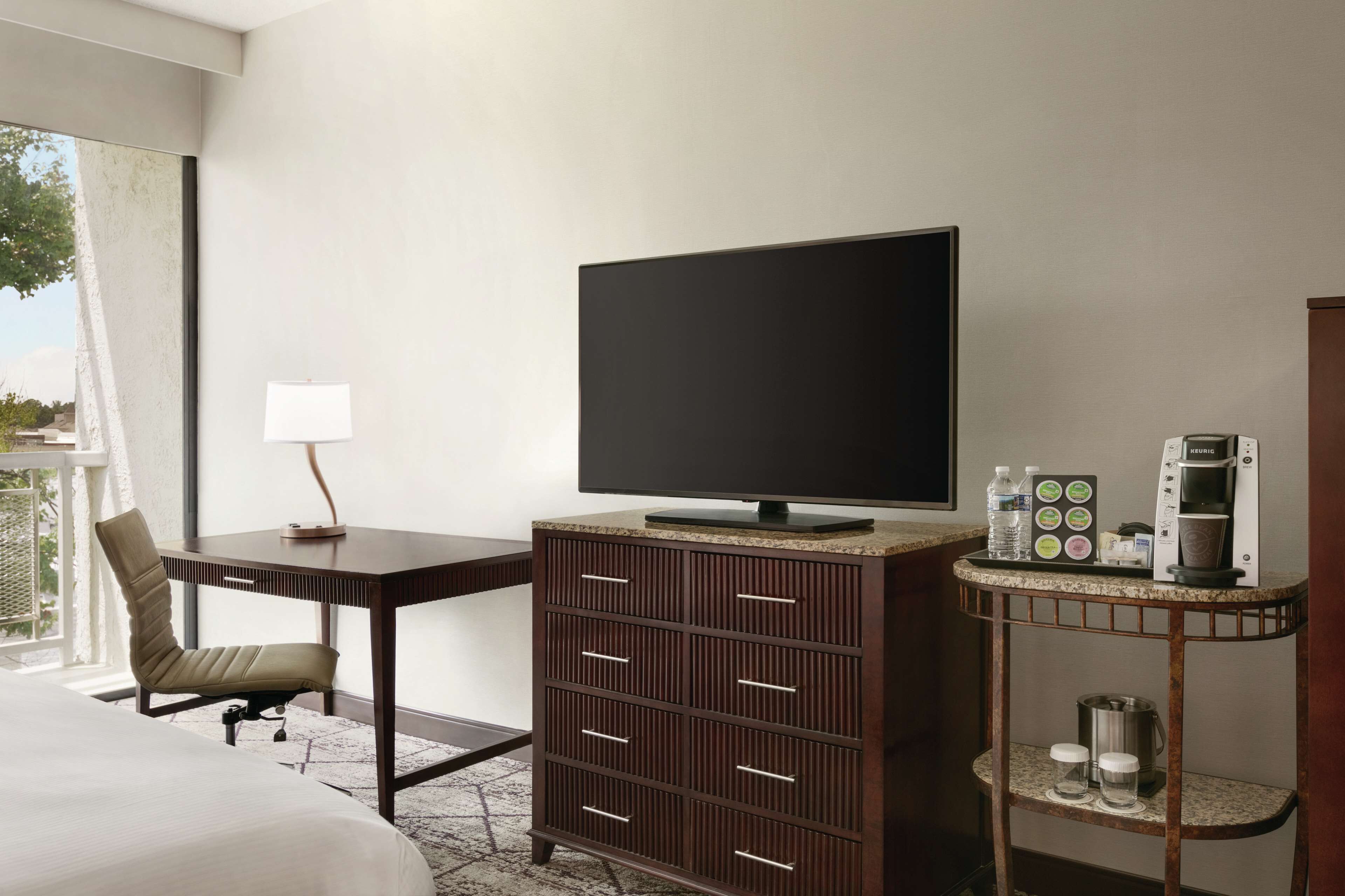 A hotel room with a bed, a desk with a TV, a coffee machine, and a minibar.