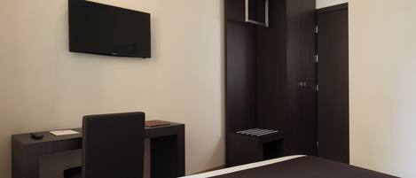 A hotel room with a bed, a desk, a chair, a TV mounted on the wall, and a wardrobe.