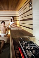 Uma sauna com ripas de madeira, uma churrasqueira a carvão e duas pessoas relaxando.