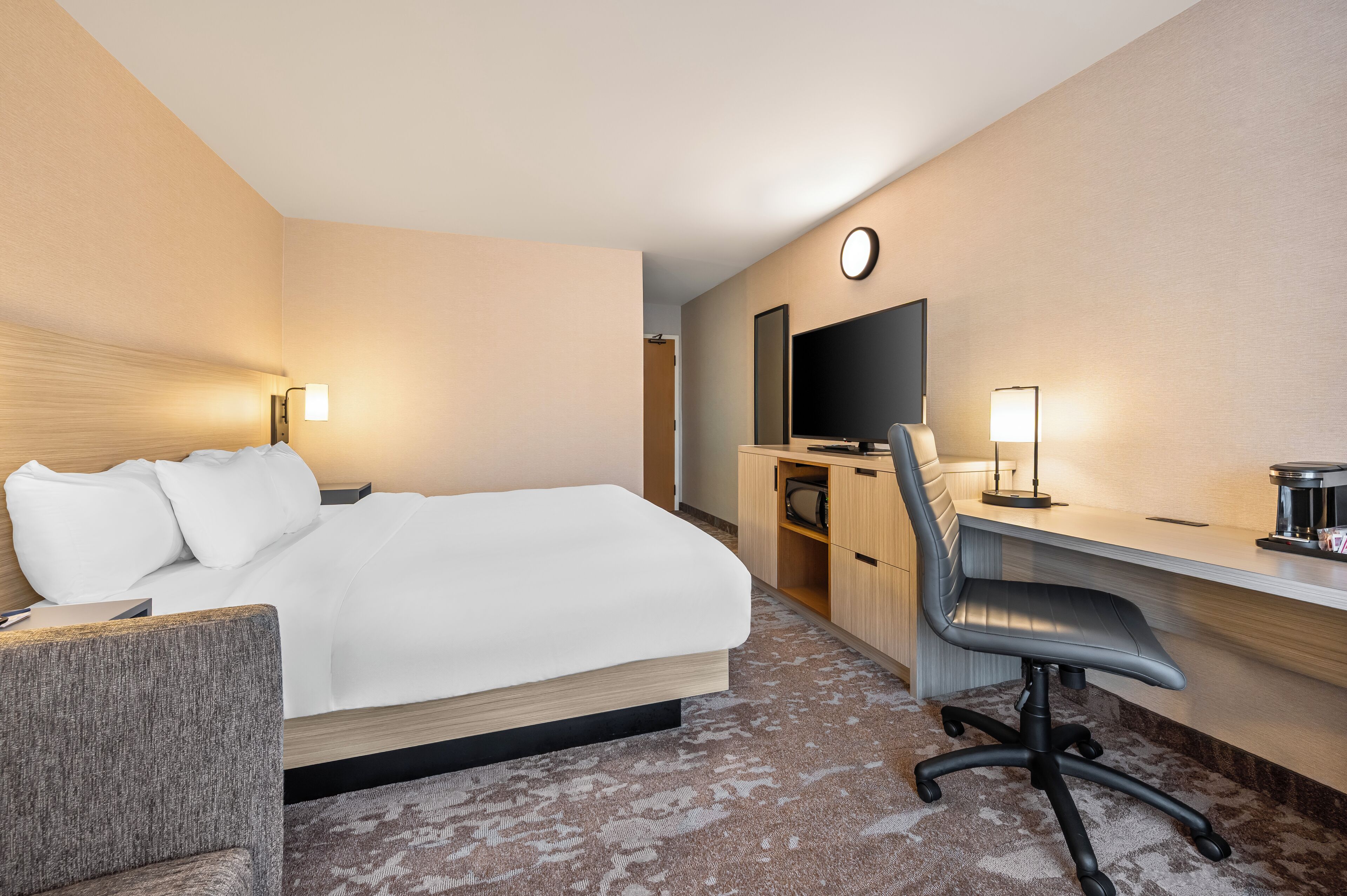 A hotel room with a bed, a desk, a chair, a television, and a clock on the wall.