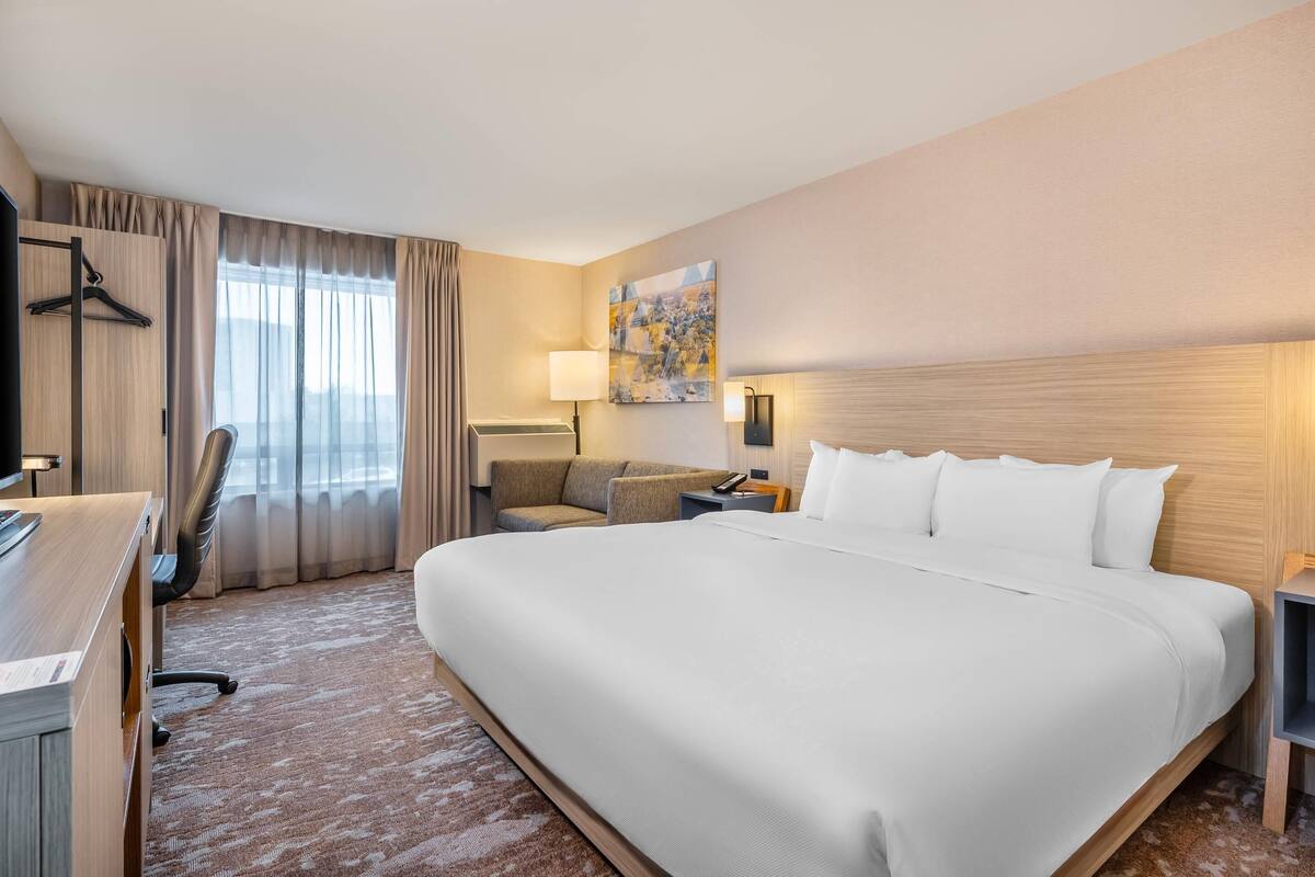 A hotel room with a large bed, a desk, a chair, a sofa, a lamp, and a painting on the wall.