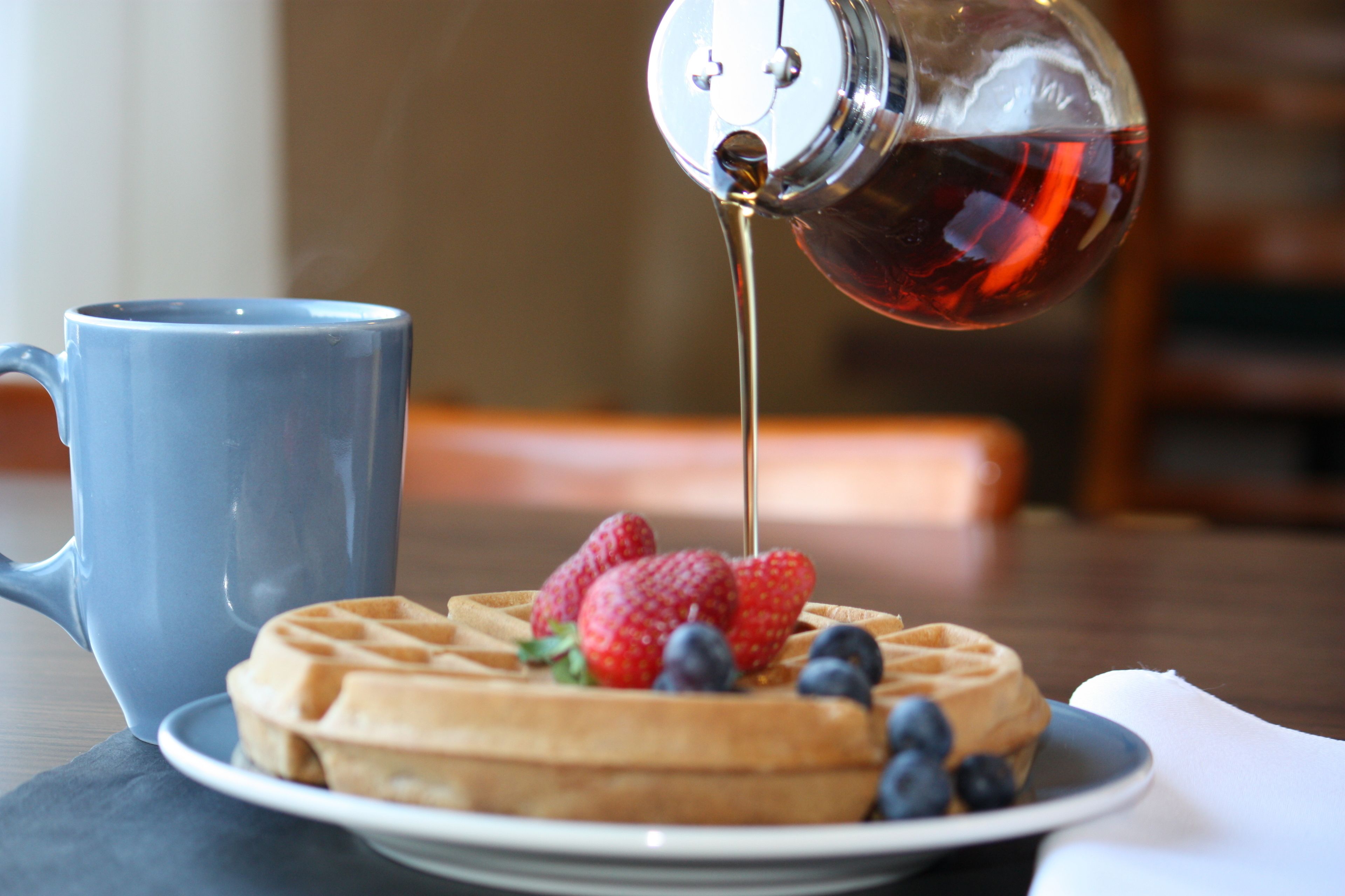 A waffle topped with strawberries and blueberries, a blue mug, and syrup being poured onto the waffle.