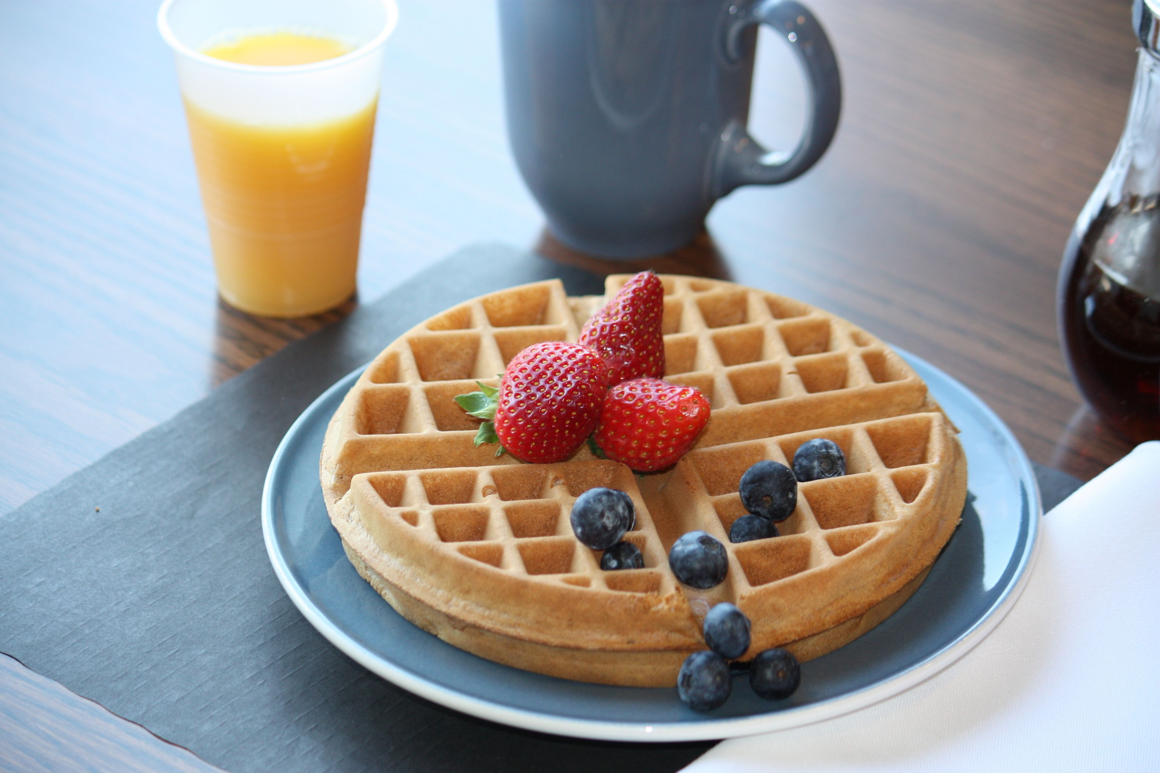 Un waffle con fresas y arándanos en un plato, un vaso de jugo de naranja y una jarrón al fondo.