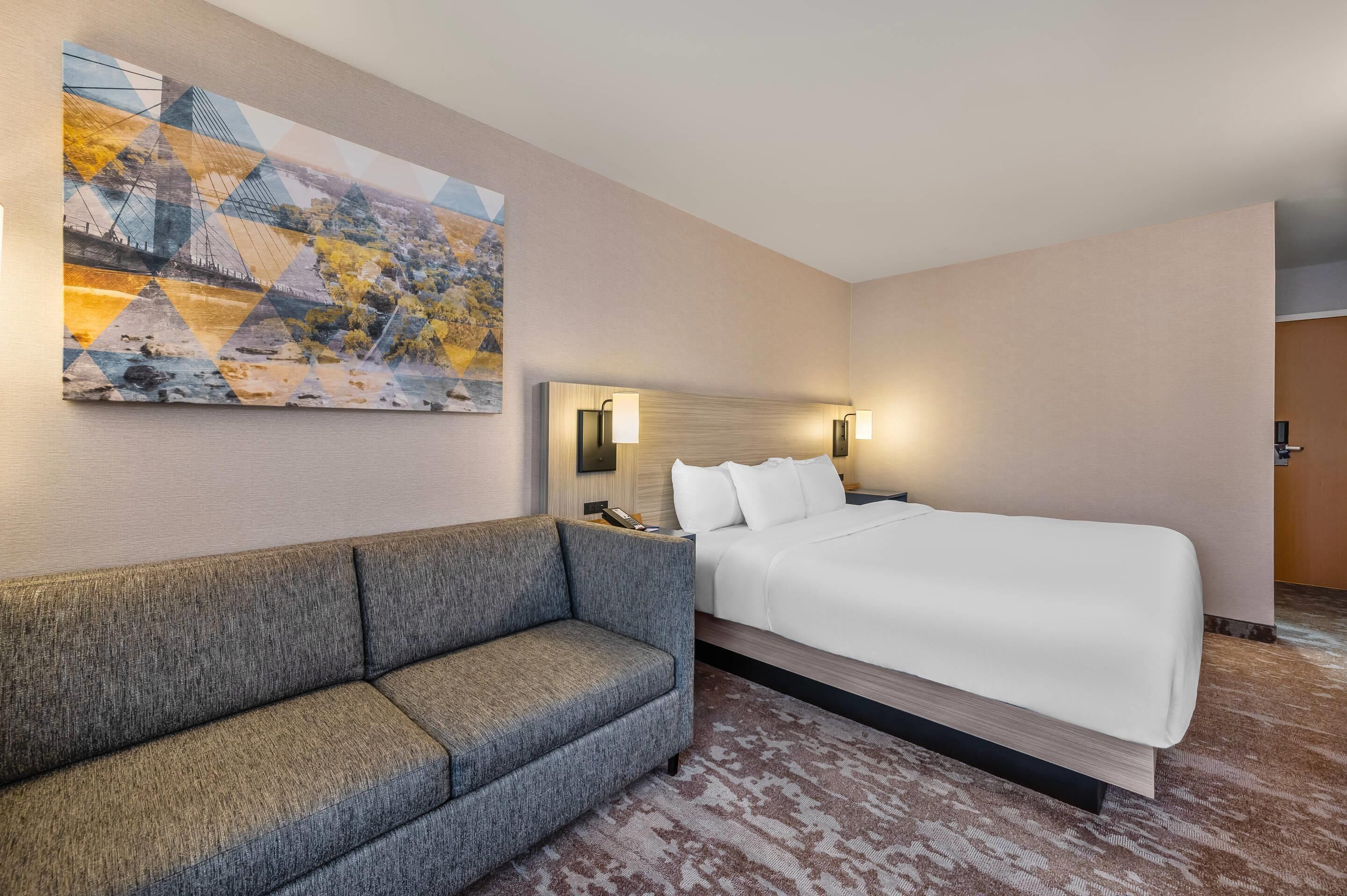 A hotel room with a large bed, a sofa, a wall-mounted artwork, and a nightstand with a lamp.