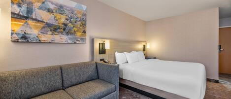 A hotel room with a large bed, a sofa, a wall-mounted artwork, and a nightstand with a lamp.
