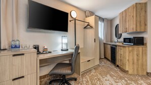 A compact hotel room with a desk, chair, flat-screen TV, bedside lamp, and a kitchenette with a microwave and refrigerator.