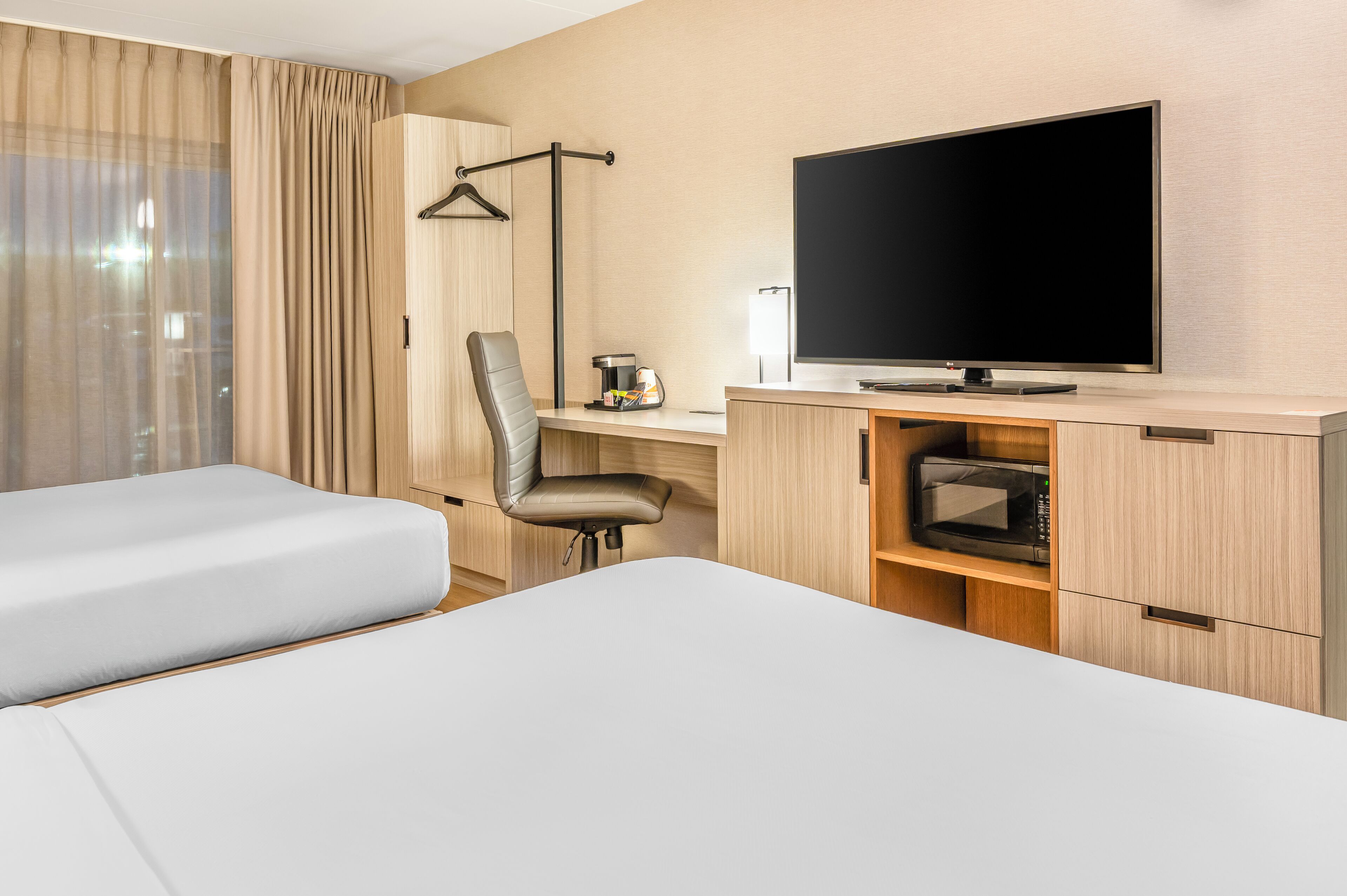 A hotel room with two beds, a desk with a flat-screen TV, a chair, a microwave, and a coat rack.